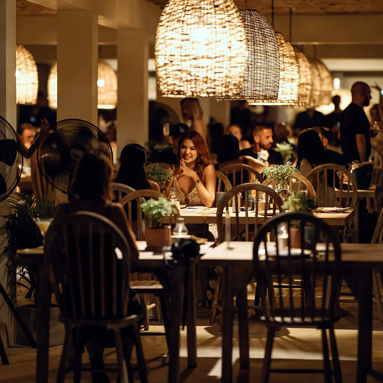 A cozy restaurant scene with warm lighting, wooden tables, and chairs. People are dining and socializing, with a woman smiling at a table in the center, surrounded by others and decorated with potted plants.