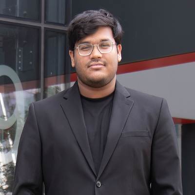 A young man with dark hair and glasses wearing a black suit jacket and shirt standing outdoors in front of a modern building.