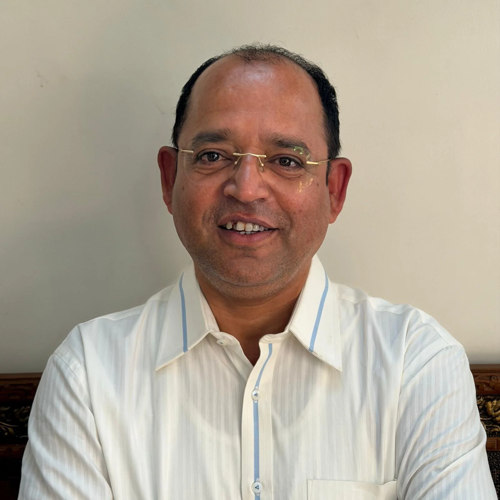 A middle-aged man with glasses and short black hair, wearing a white collared shirt, smiling and sitting in front of a plain light-colored wall.