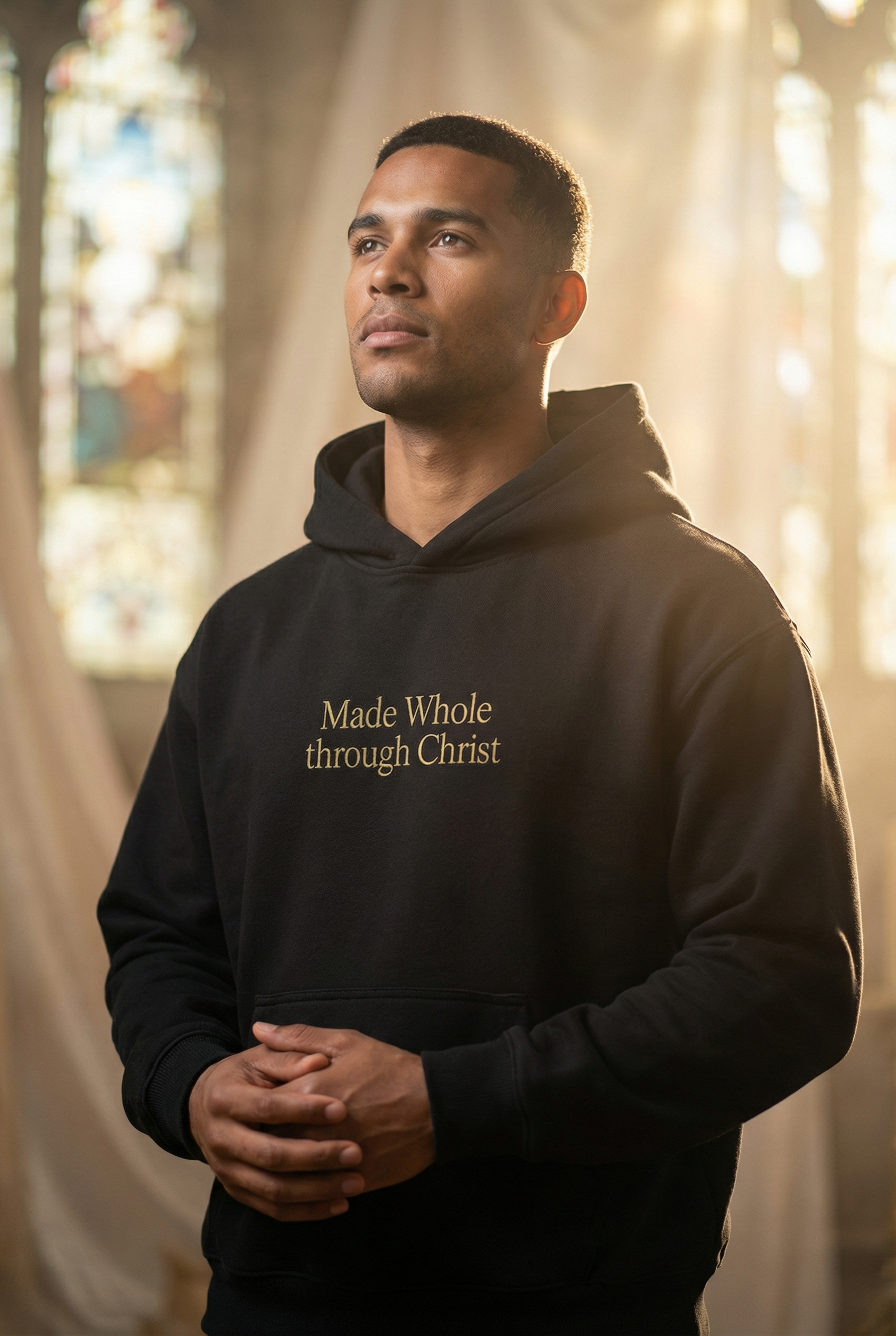 A young man standing inside a church with stained glass windows, wearing a black hoodie with the words "Made Whole through Christ"  on the front, looking contemplative.