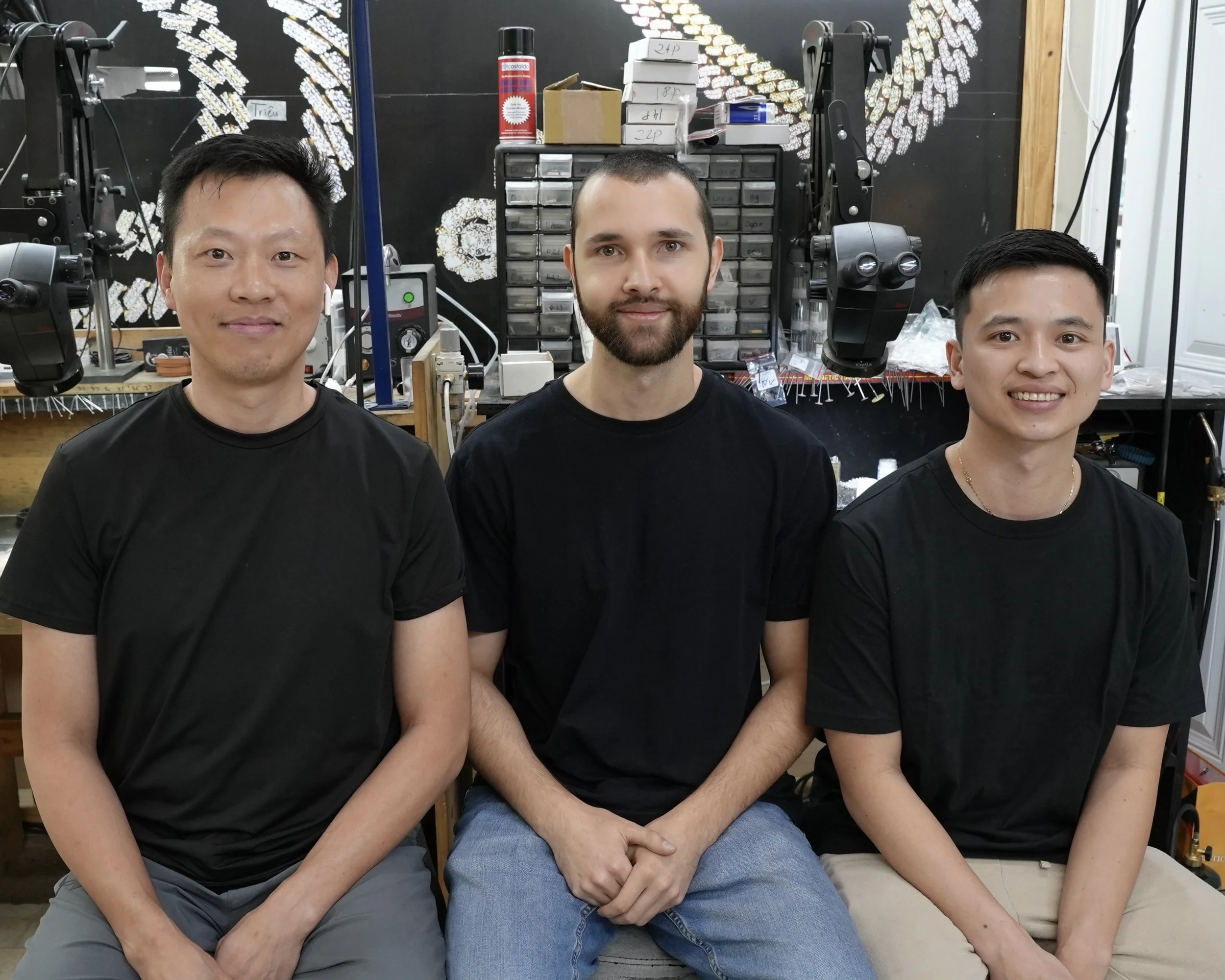 Three men sitting side by side indoors, with a workshop or laboratory in the background, including microscopes, drawers, and scientific equipment.