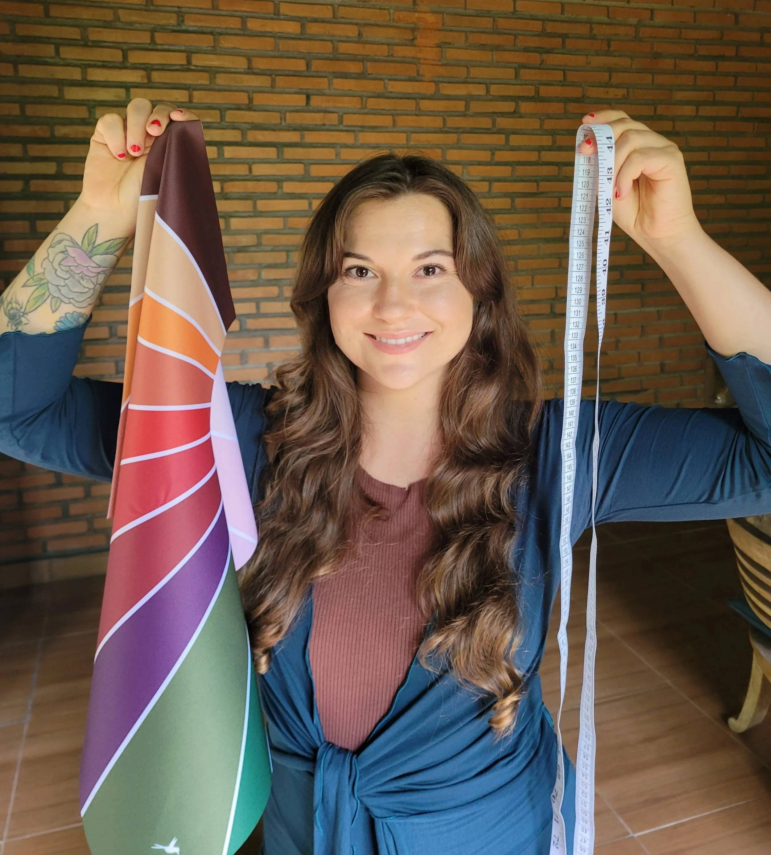 A woman with long, wavy brown hair smiling at the camera, holding a colorful umbrella in her left hand and a measuring tape in her right hand. She is standing indoors against a brick wall.