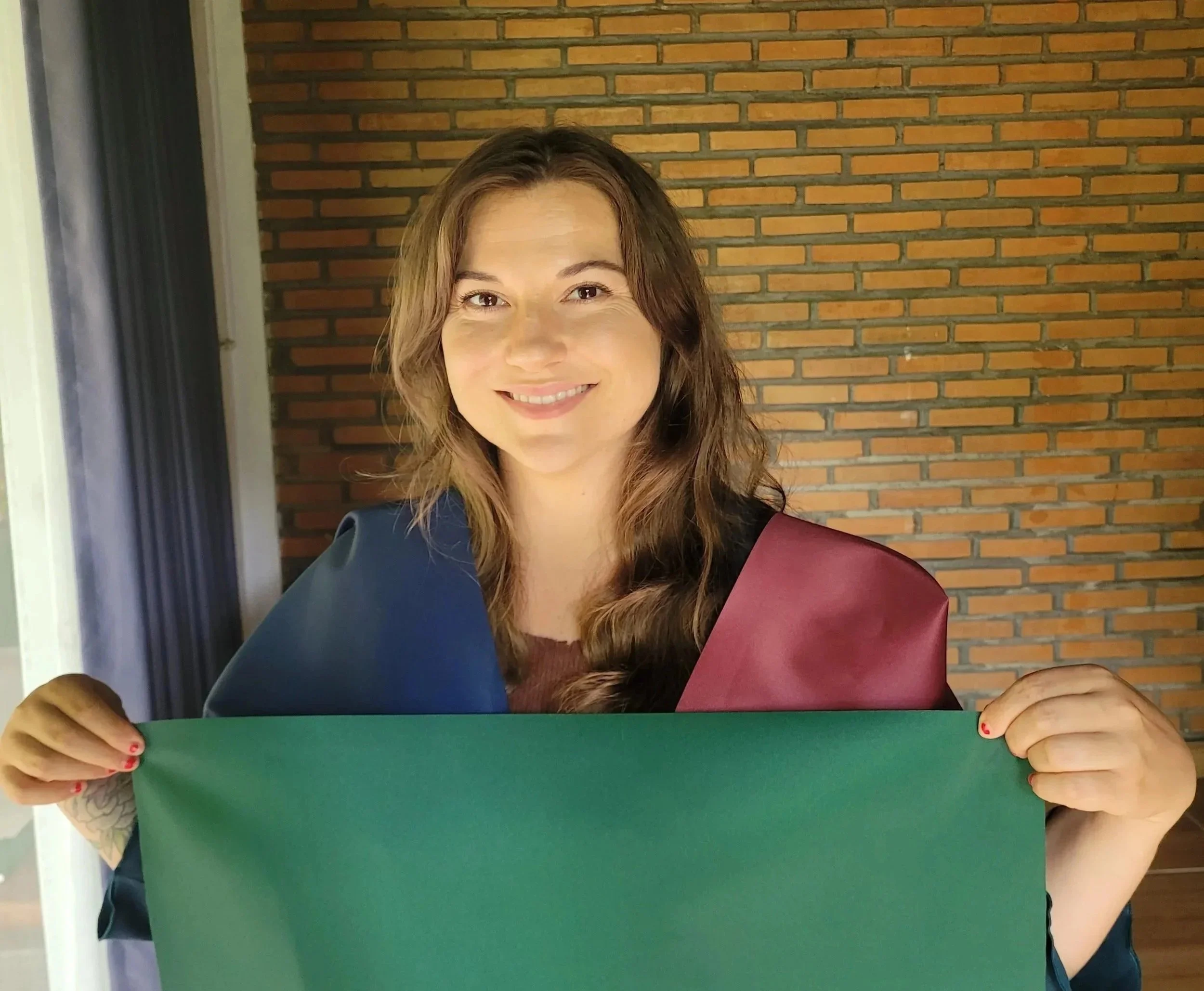 Woman smiling, holding a green card in front of her, with a brick wall background.