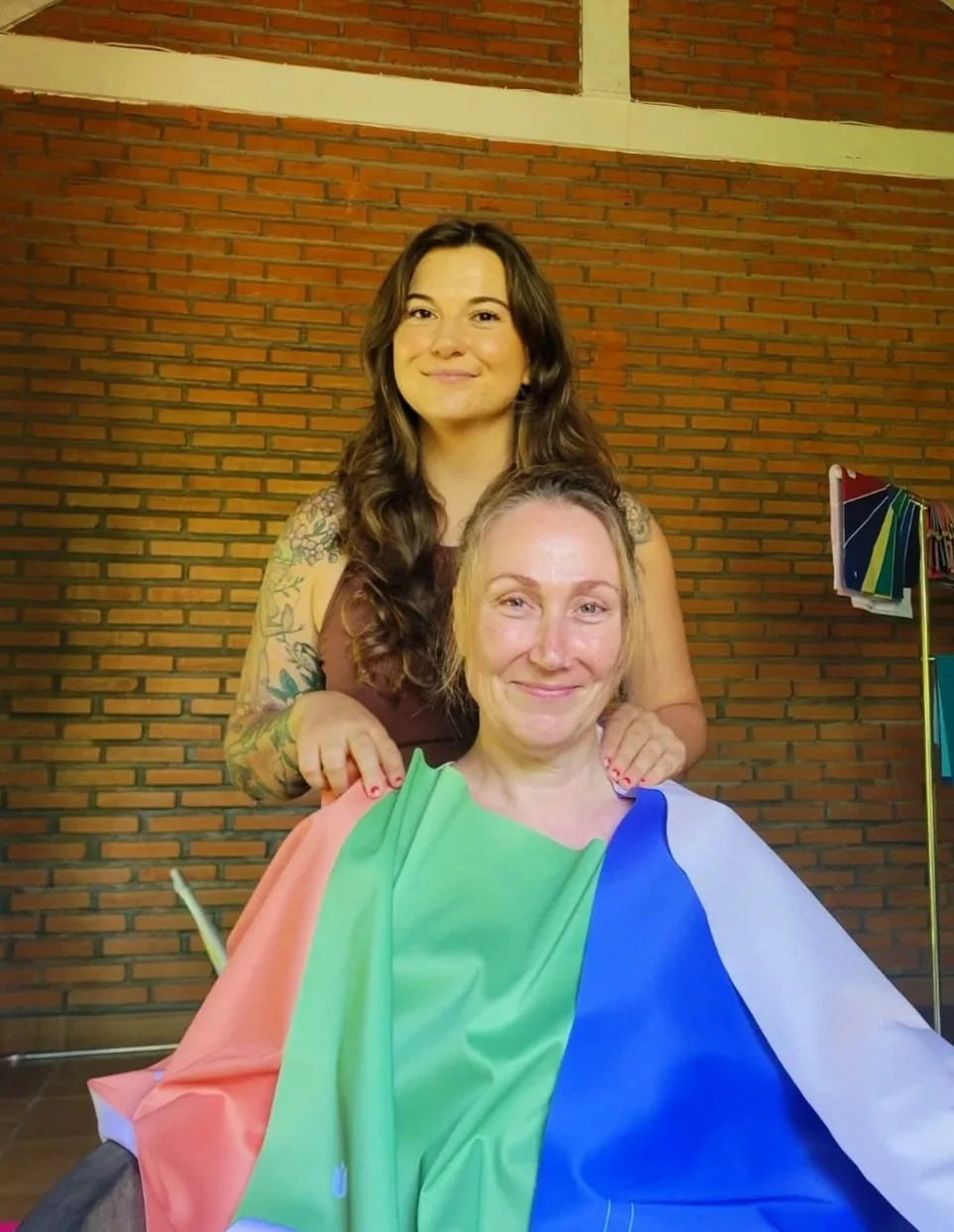 Two smiling women, one standing behind the other, indoors against a brick wall. The seated woman has a rainbow-colored fabric draped over her shoulders, resembling a flag.