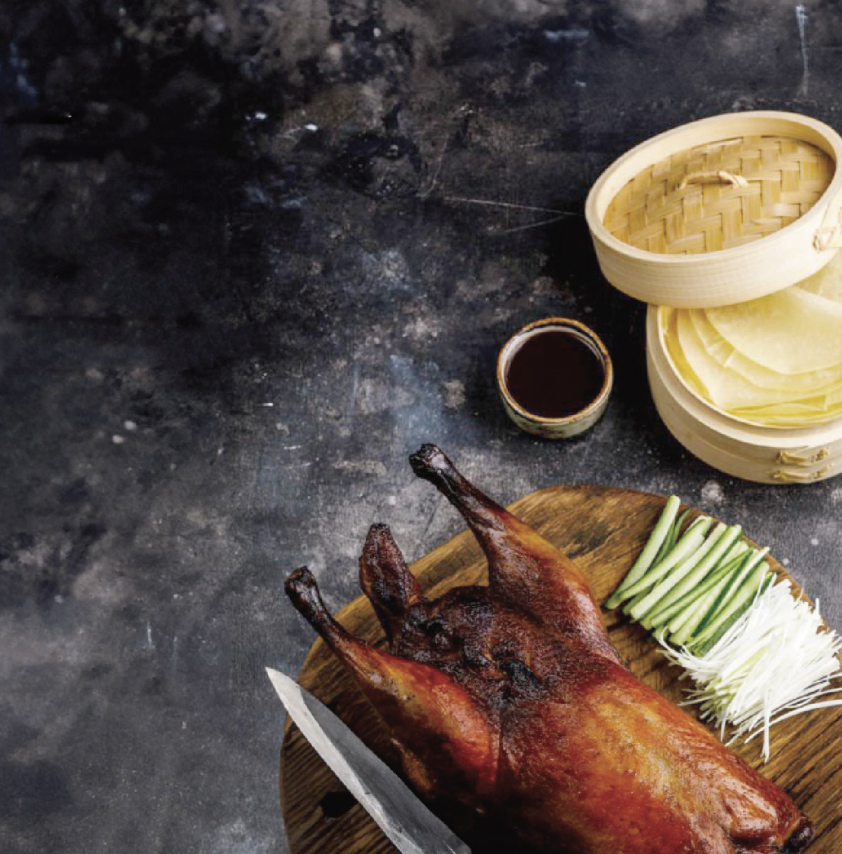 Roasted Peking duck with sliced cucumber and scallions on a wooden board, side of hoisin sauce in a small cup, and bamboo steamers with thin pancakes in the background.