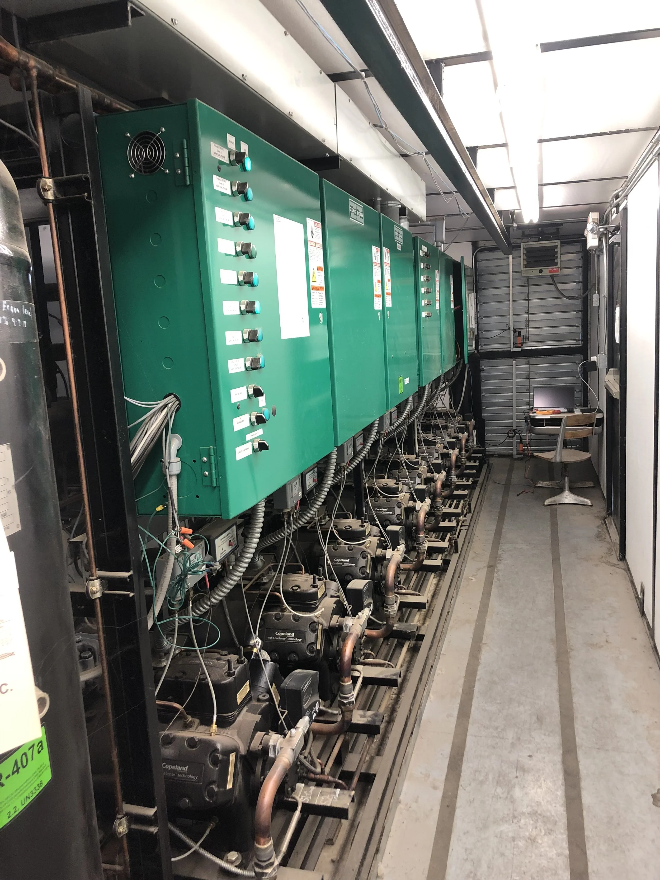 Equipment room with large green electrical control panels, numerous cables and pipes, and a small desk with a chair in the corner.