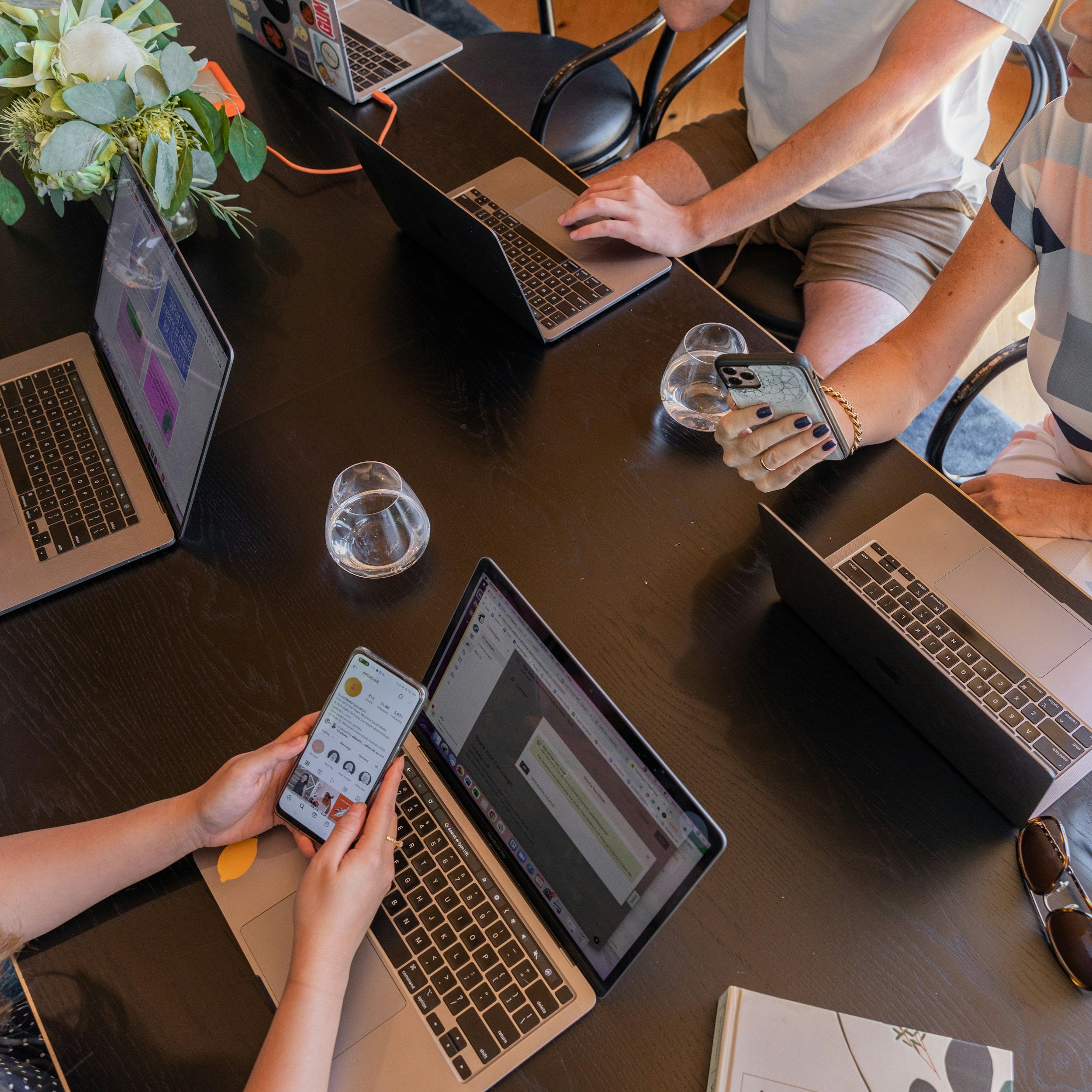 People sitting around a black table with laptops, smartphones, glasses of water, and a flower arrangement, engaged in work or conversation.