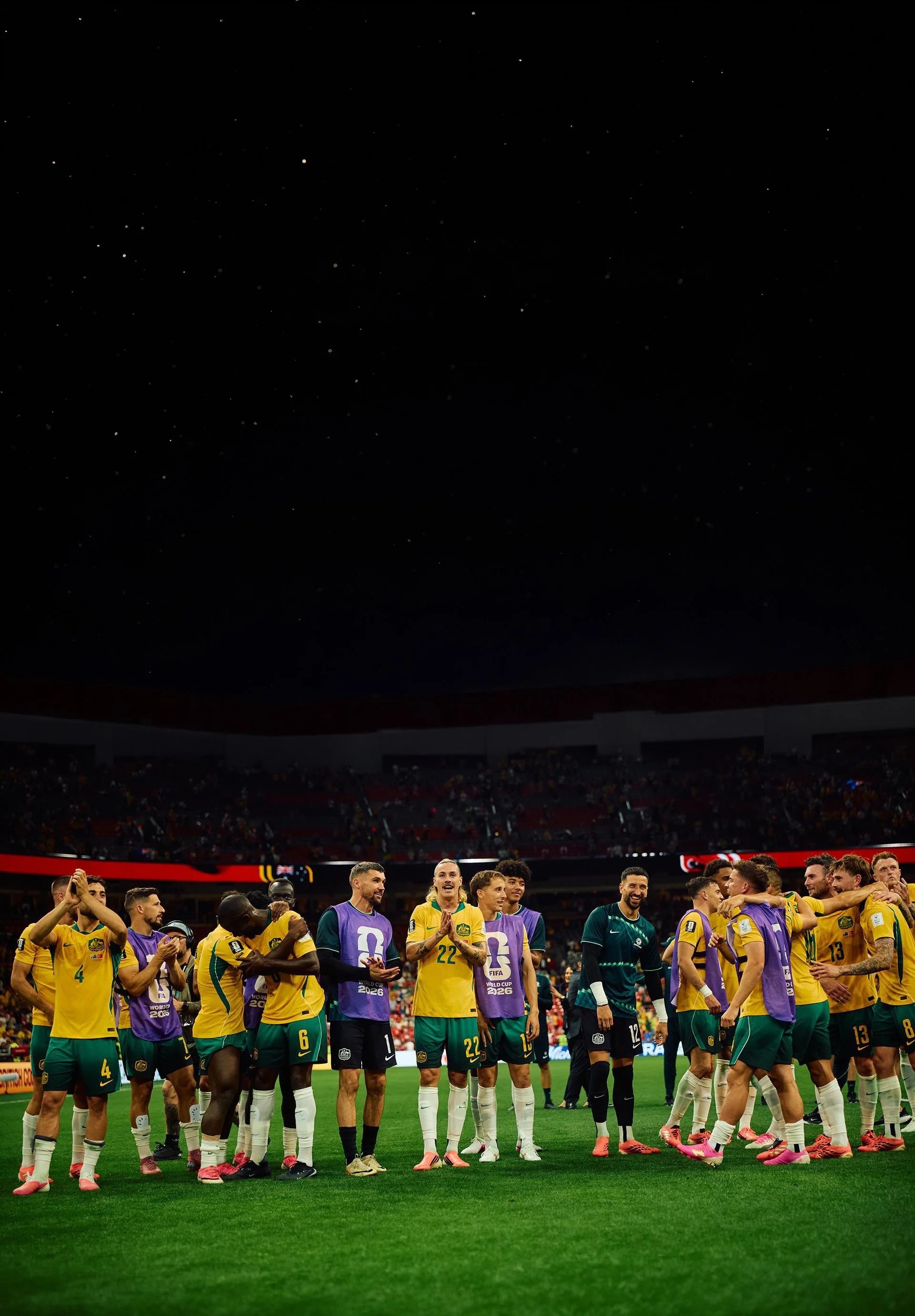 Soccer players standing in line on the field at a stadium during a match, with some players celebrating or talking to each other, and a large crowd in the stands.