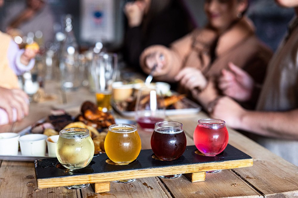 A flight of four colorful beers on a wooden paddle in front of people enjoying a meal at a restaurant. The beers are pale yellow, amber, dark reddish, and bright pink.