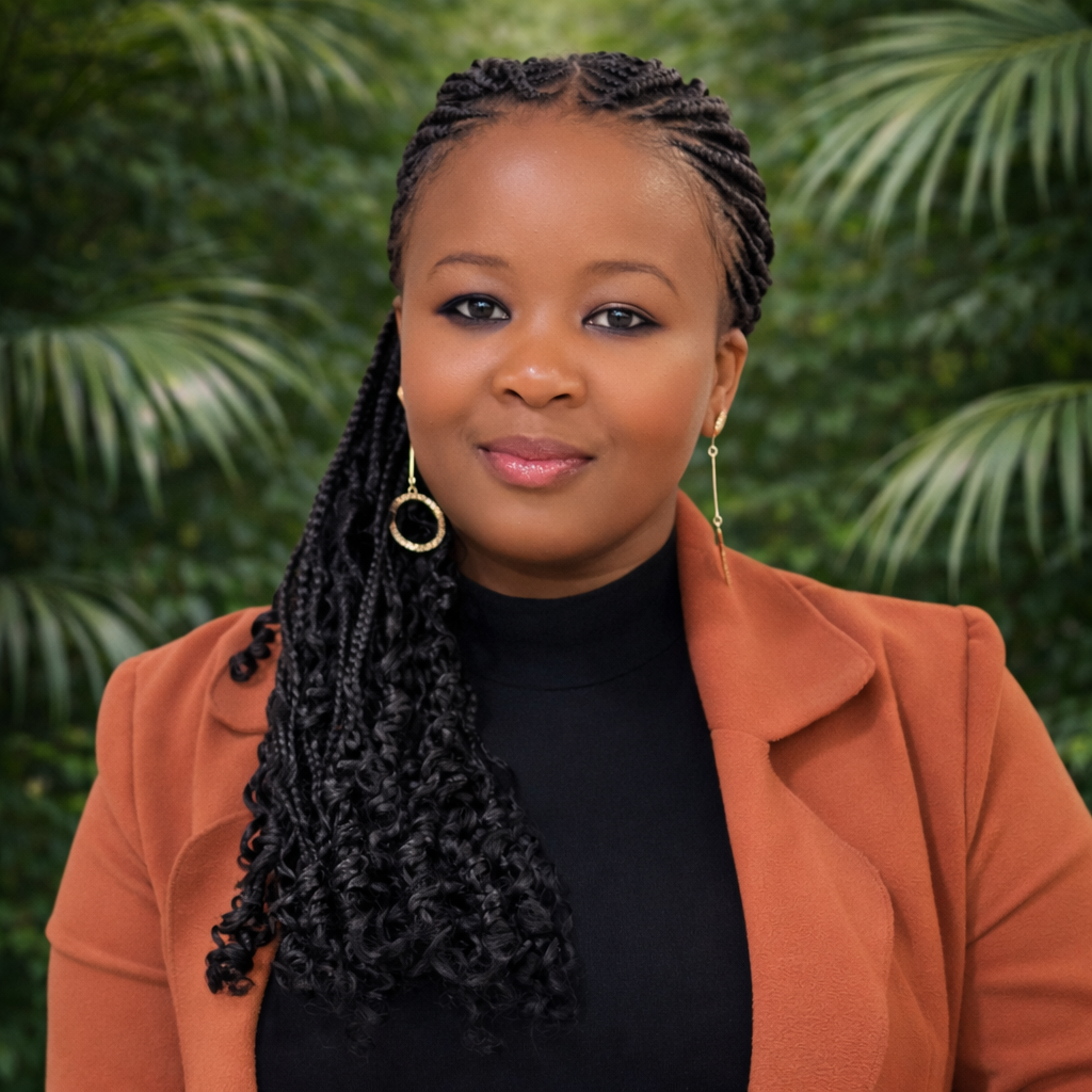 A woman with styled braids, wearing a brown blazer and black top, standing outdoors with green foliage in the background.