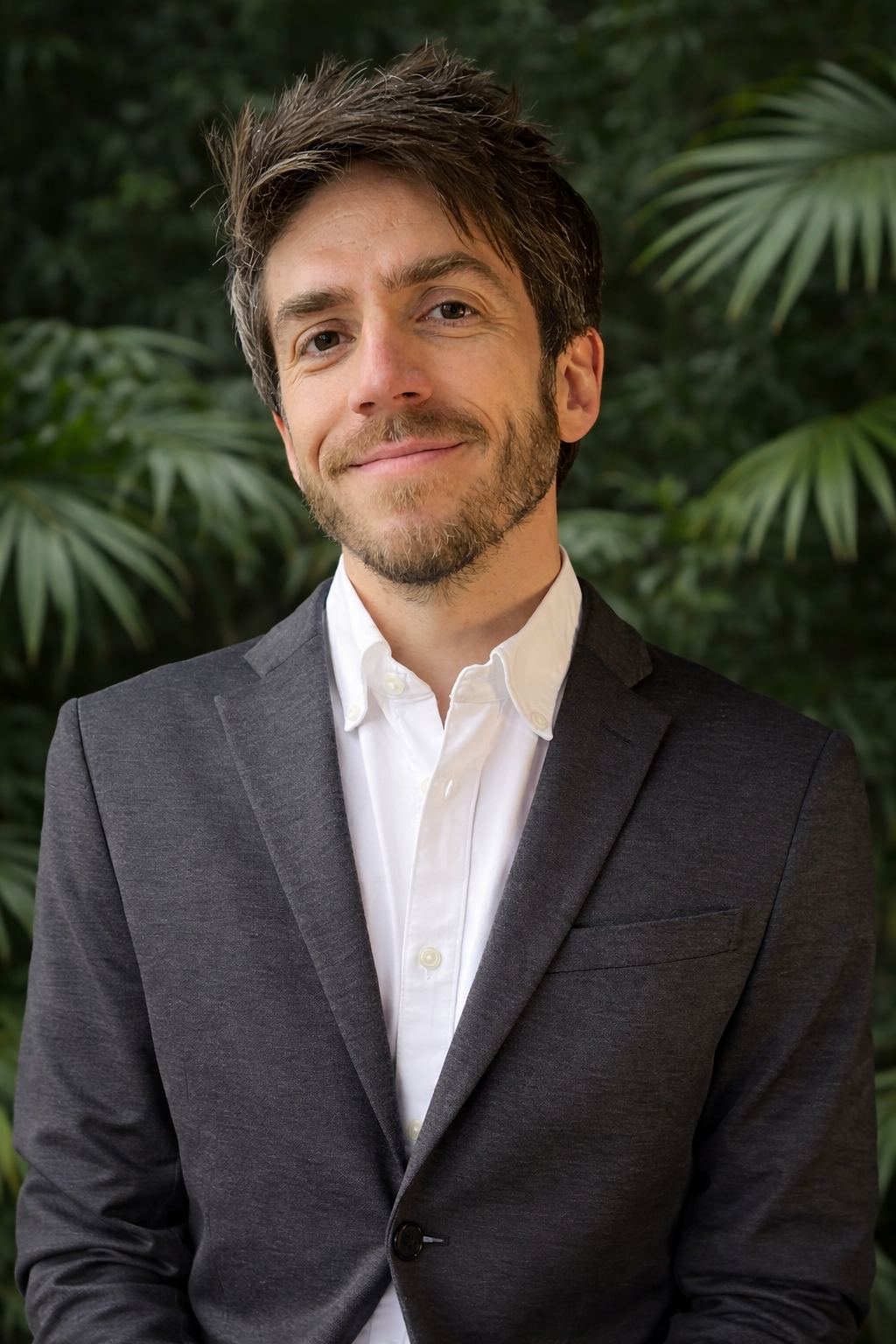 A smiling man in a dark suit jacket and white shirt standing outdoors with green tropical plants in the background.