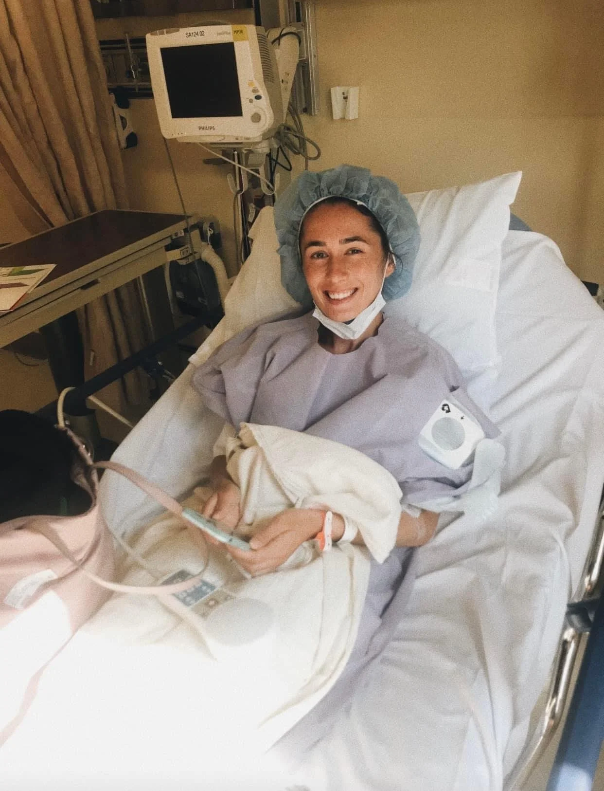 Smiling woman in hospital bed wearing a hospital gown and hair cap,  with medical monitors and equipment around her.