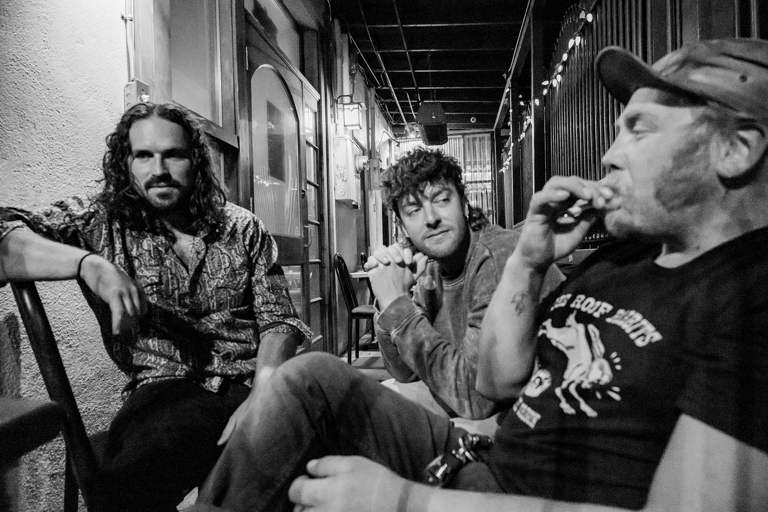 Three men sitting on chairs outside a cafe or bar, having drinks and conversing. One man is on the right drinking from a glass, wearing a cap and a graphic T-shirt. The other two men are seated facing toward the camera, one with long curly hair and the other with short curly hair