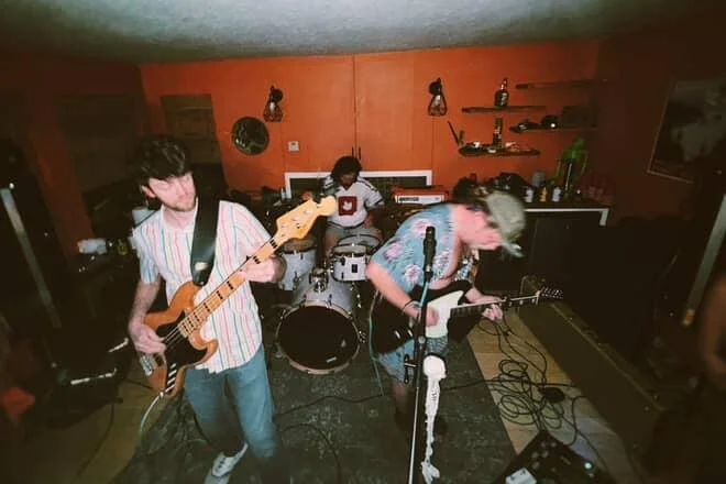 Three musicians playing guitars and drums in a cozy indoor setting.
