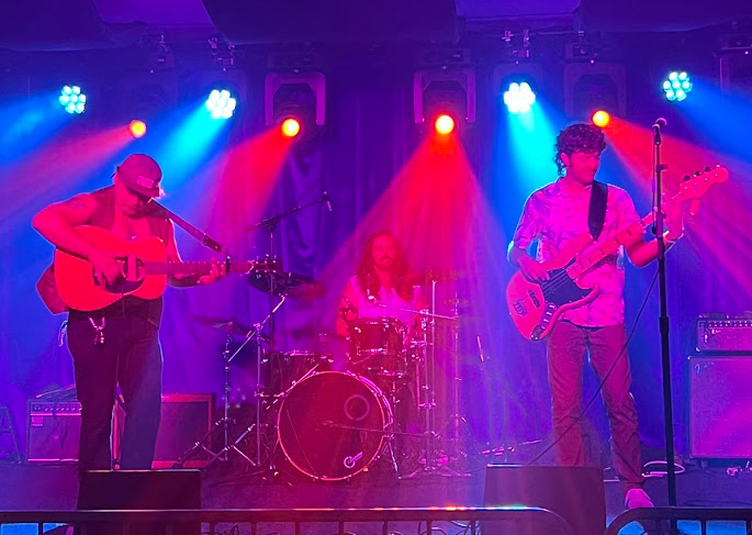 A band performing on stage with colorful lighting, featuring a guitarist, a drummer, and a bassist.