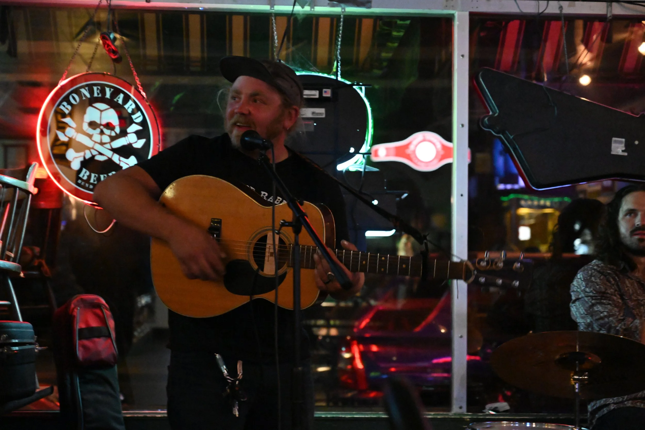 A man with a beard and wearing a cap is playing an acoustic guitar and singing into a microphone at a bar or music venue. Neon signs and dim lighting create a lively atmosphere.