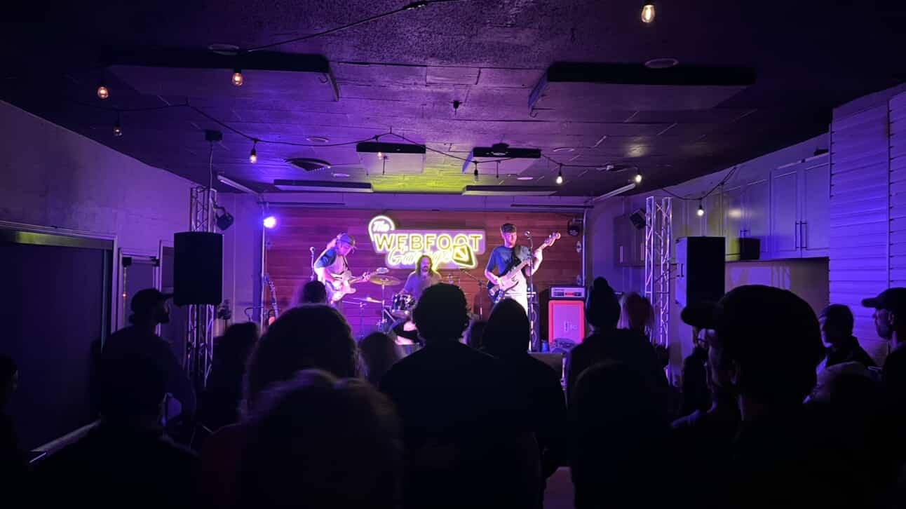Live band performing on stage at a small indoor concert venue with an audience watching, neon sign behind the band reads 'WEBFOG'
