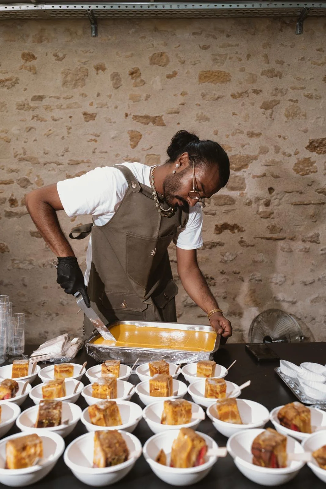 A man wearing glasses inspects a tray of food while preparing desserts at a table filled with square portions of cake in small white bowls, against a stone wall background.