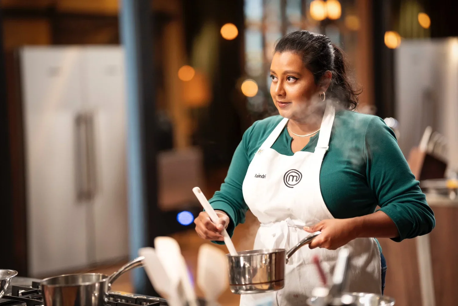 A woman wearing a white apron with the 'MasterChef' logo and the name 'Saindi' on it, stirring a pot in a professional kitchen. She has dark hair pulled back and is wearing a green long-sleeve shirt.