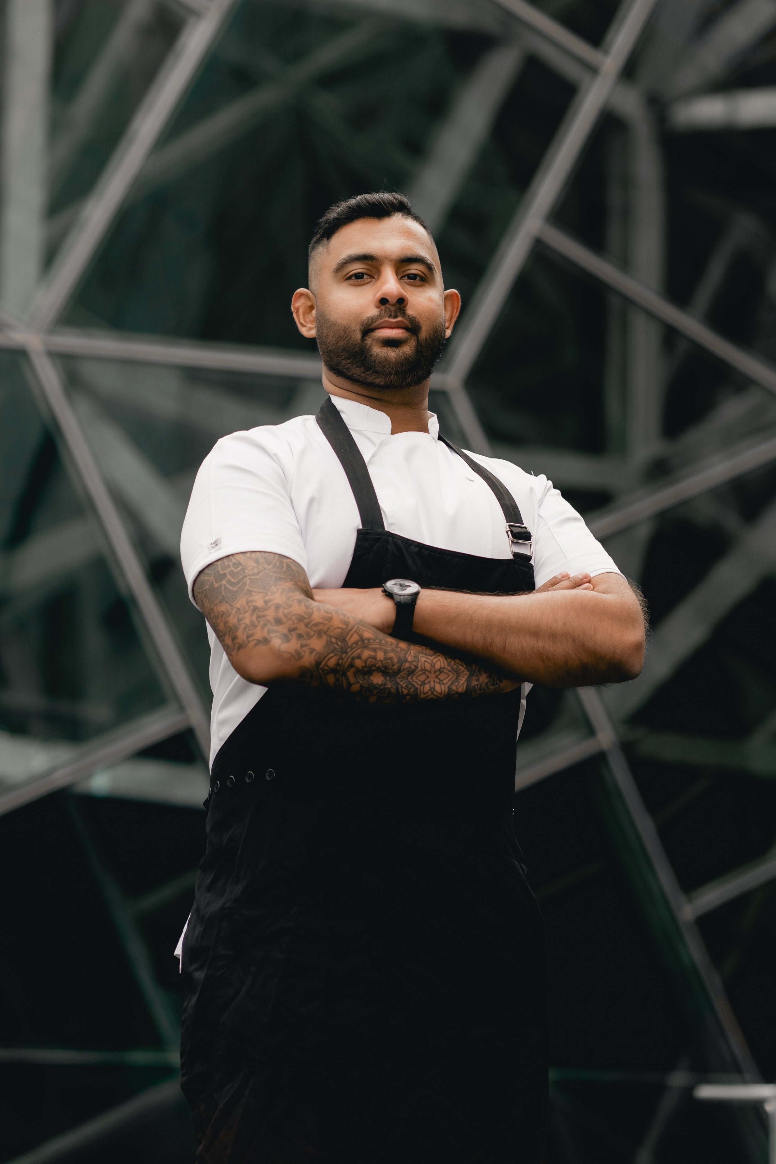 A confident male chef with tattoos on his left arm, standing with arms crossed in front of a reflective, geometric glass building.