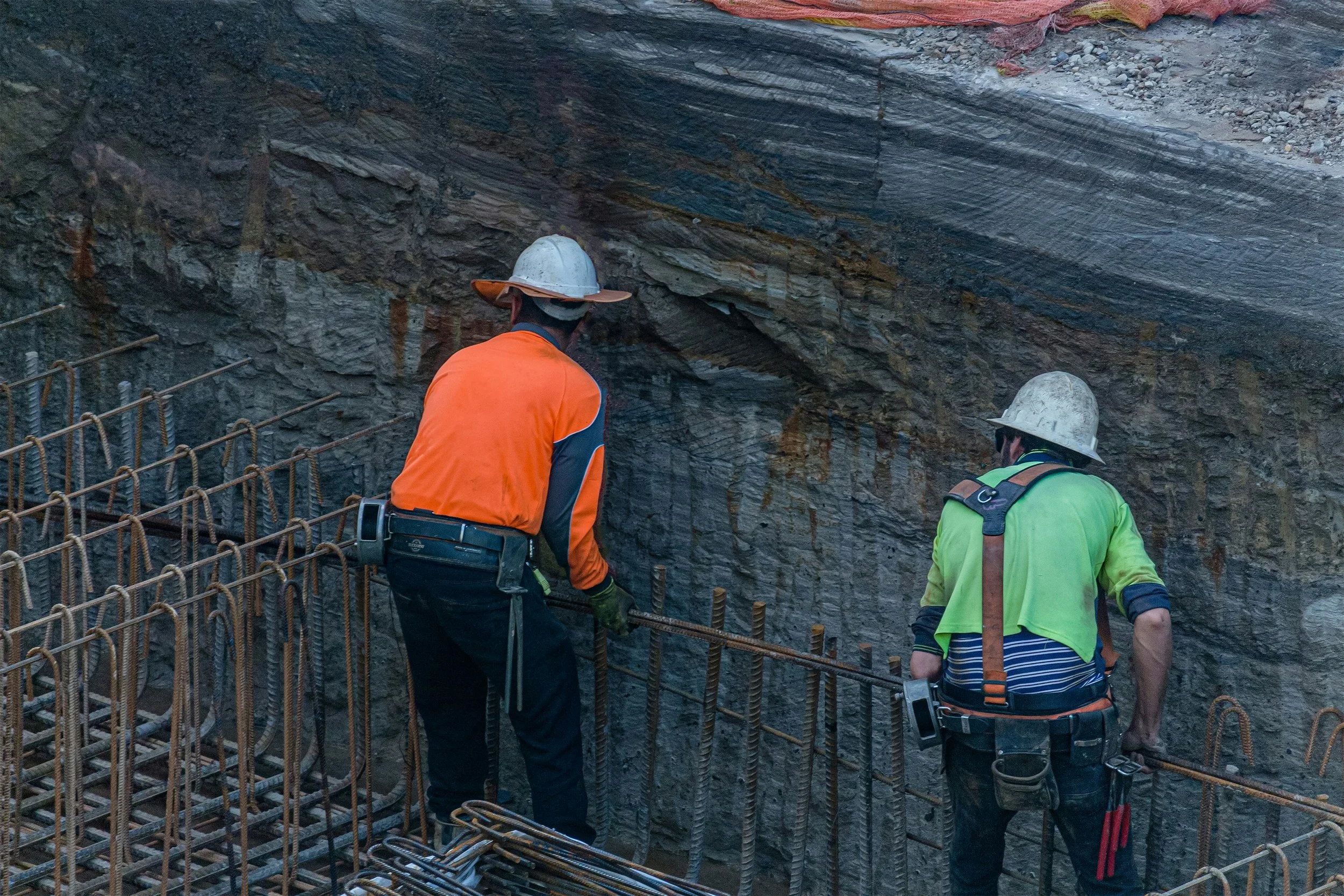 Two construction workers wearing safety helmets and high-visibility vests working on steel reinforcement bars inside a concrete structure at a construction site.