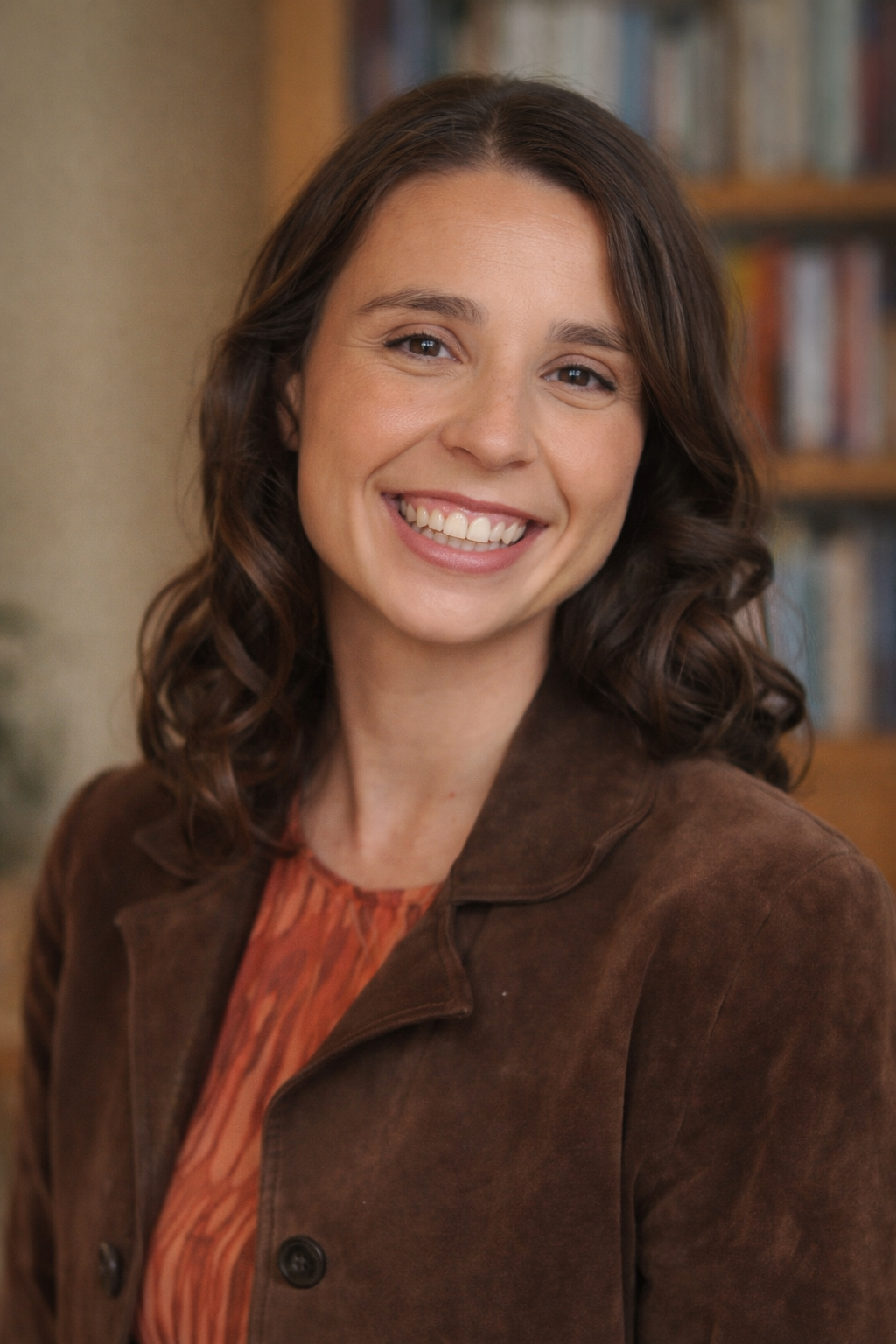 A woman with wavy brown hair, smiling, wearing a brown jacket and an orange patterned blouse, standing indoors in front of a bookshelf.