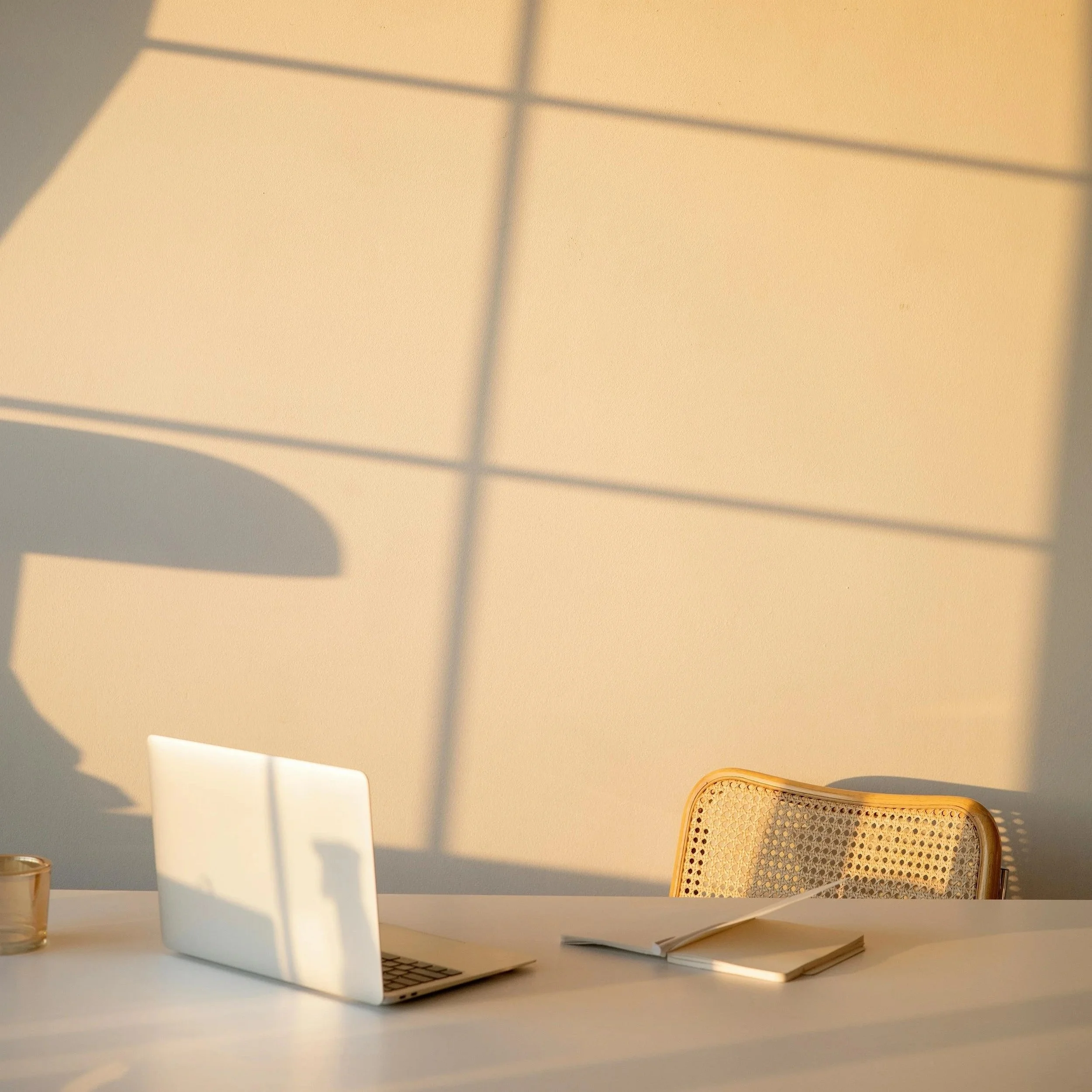 Sunlight casting shadows of window panes on a beige wall behind a white desk. On the desk, there is a closed silver laptop, a small empty glass container, an open notebook with a pen, and a rattan chair with a perforated backrest.