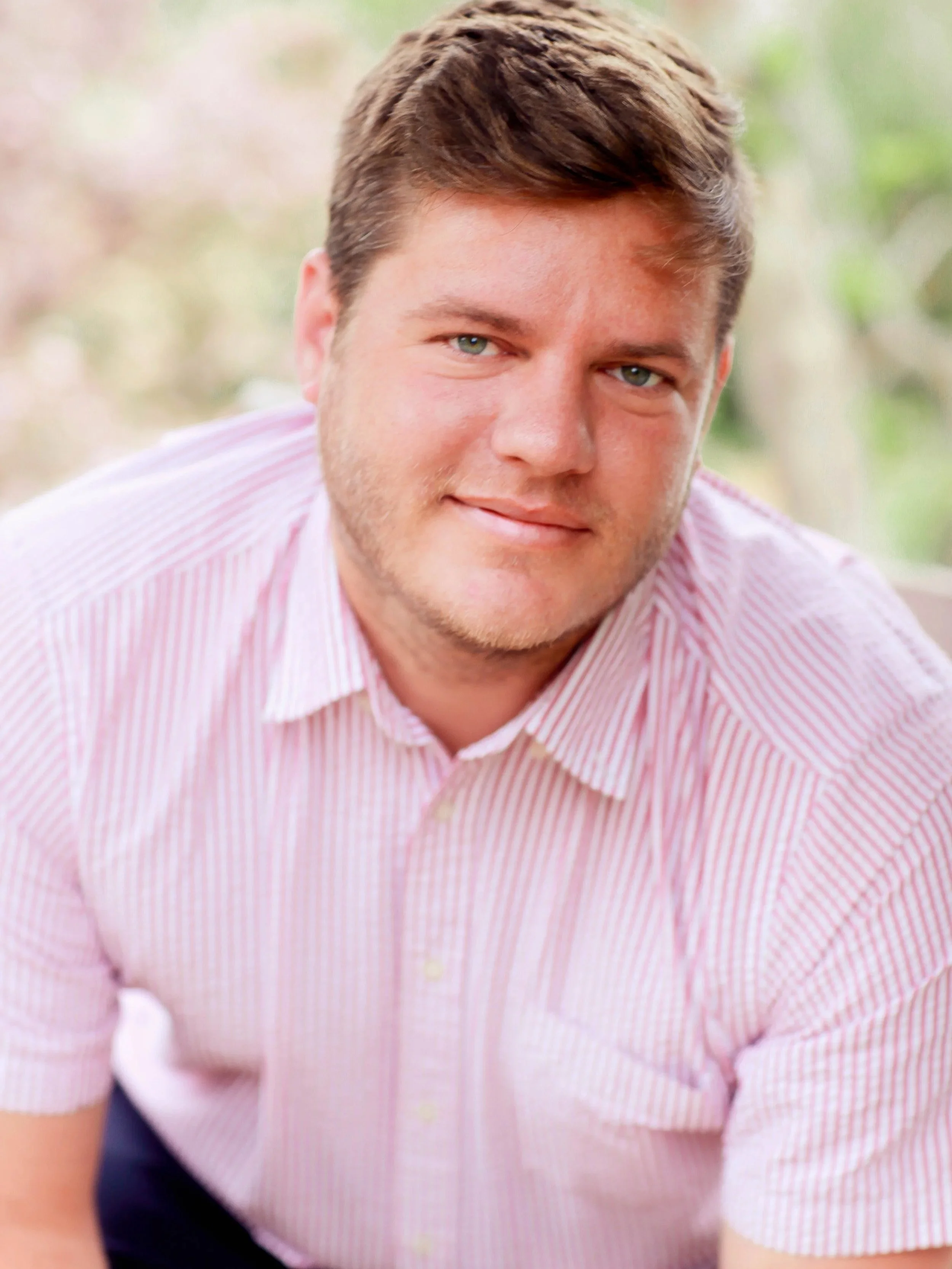 Portrait of a young man with light brown hair and a light beard, wearing a pink and white striped shirt, smiling softly outdoors with blurred greenery in the background.
