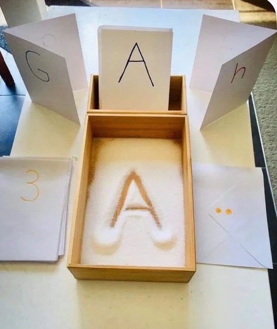 Educational alphabet learning activity with cards featuring letters G, A, H, and a number 3, with a large letter A traced in sand in a wooden box.