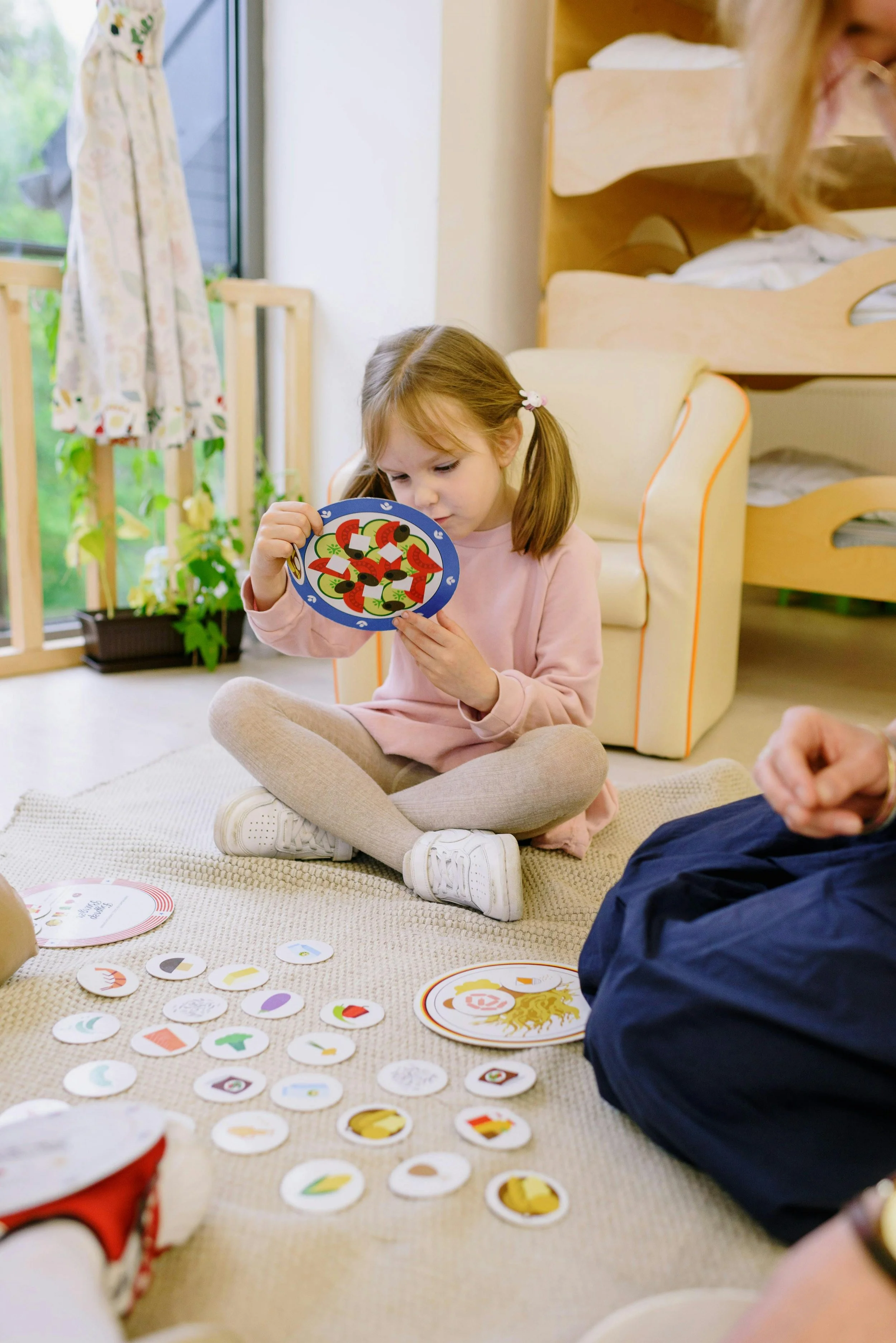 Girl sitting on beige carpet, playing a colorful card game with circular cards, near bed with bunk beds in a bright room with curtains and plants.