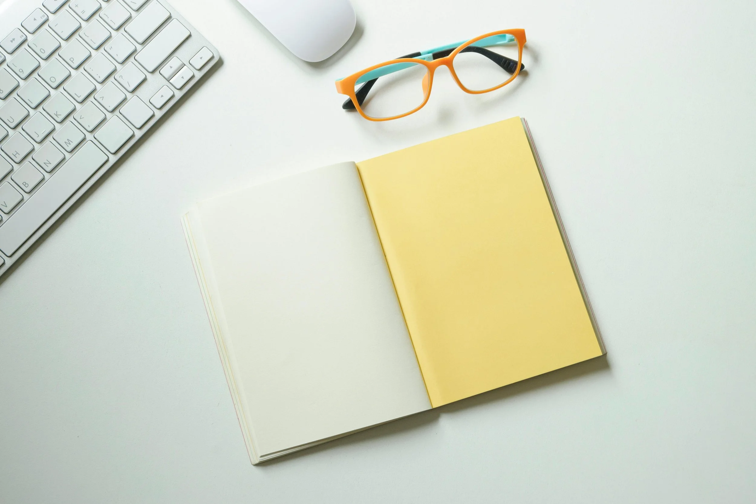 A white desk with a keyboard, a mouse, a pair of orange and teal eyeglasses, and an open notebook with yellow pages.
