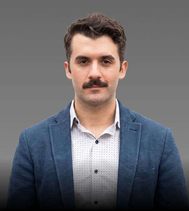 A man with dark hair and a mustache wearing a blue blazer and a white collared shirt with small dark polka dots on a plain gray background.