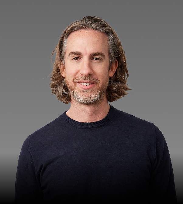 A man with shoulder-length gray hair and a beard, smiling, wearing a dark navy sweater, standing against a gray backdrop.