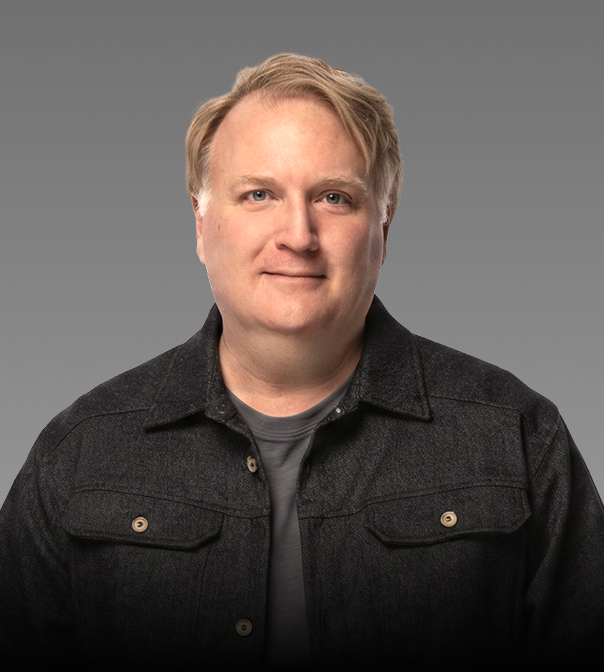 Portrait of a man with blonde hair wearing a dark jacket and gray t-shirt, against a gray background.
