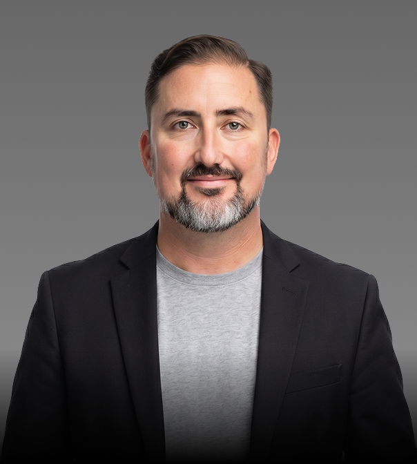 A man with dark hair and a beard, wearing a gray T-shirt and black blazer, smiling against a plain gray background.