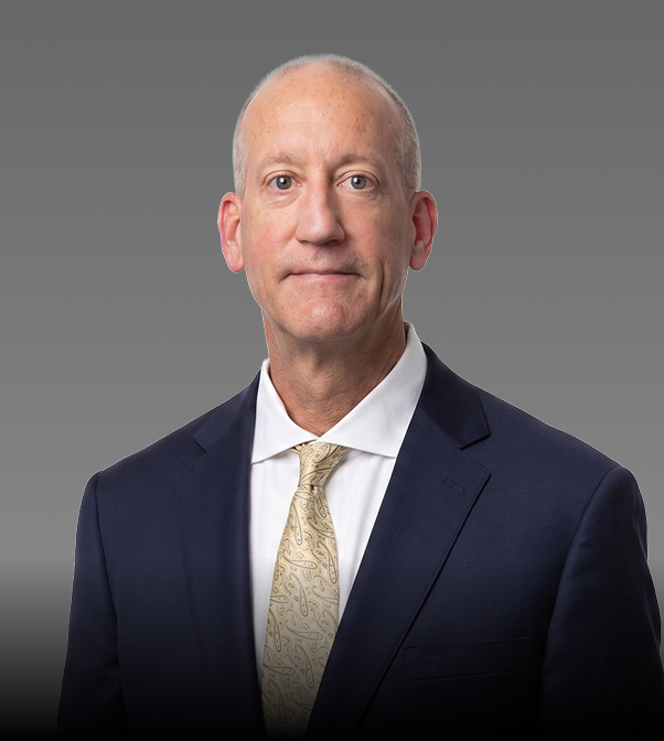 A middle-aged man in a navy blue suit, white shirt, and light yellow tie standing against a gray background.