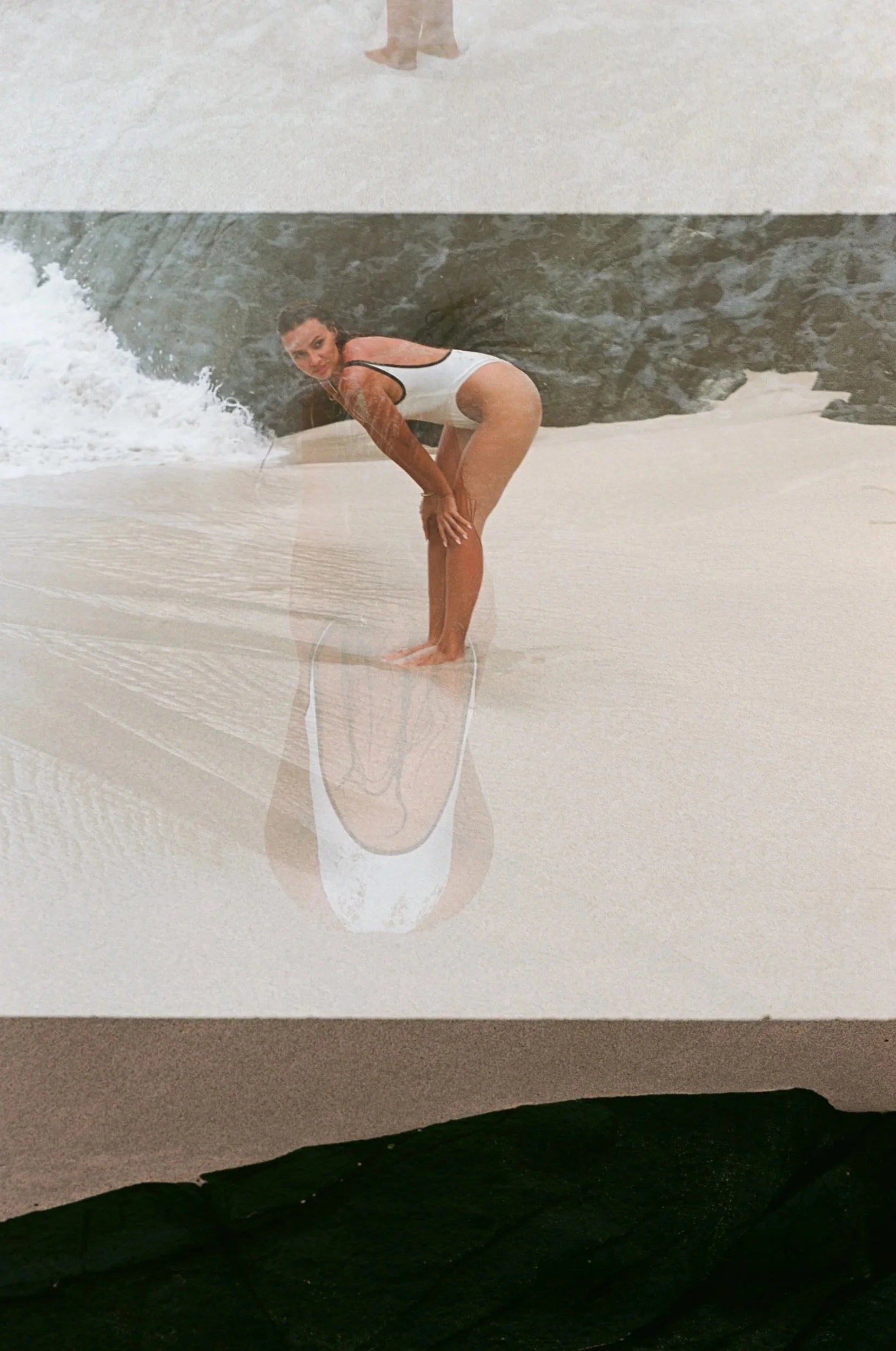 A woman in a white swimsuit bending over on a sandy beach, with her reflection visible in a puddle of water.