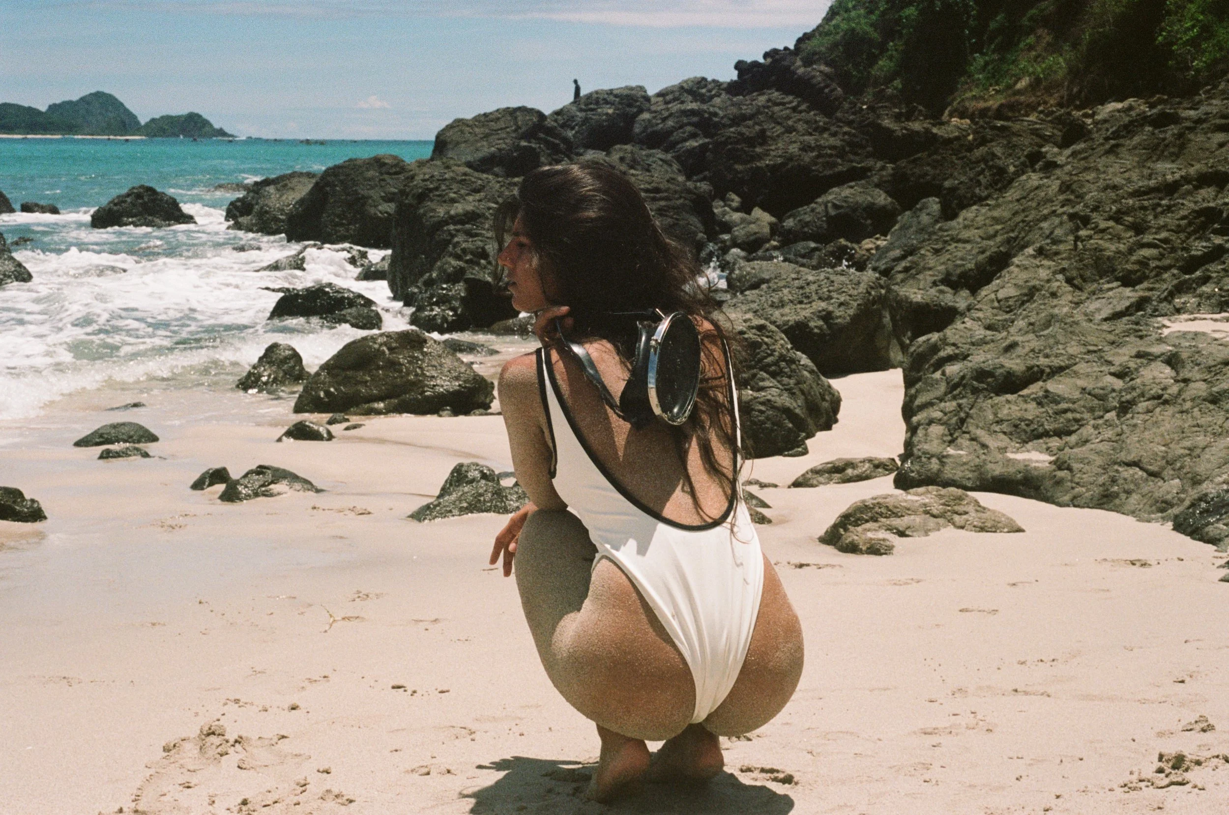 A woman with long dark hair squatting on a sandy beach near rocks, with ocean waves and a small island in the background, wearing a white swimsuit and scuba diving mask.