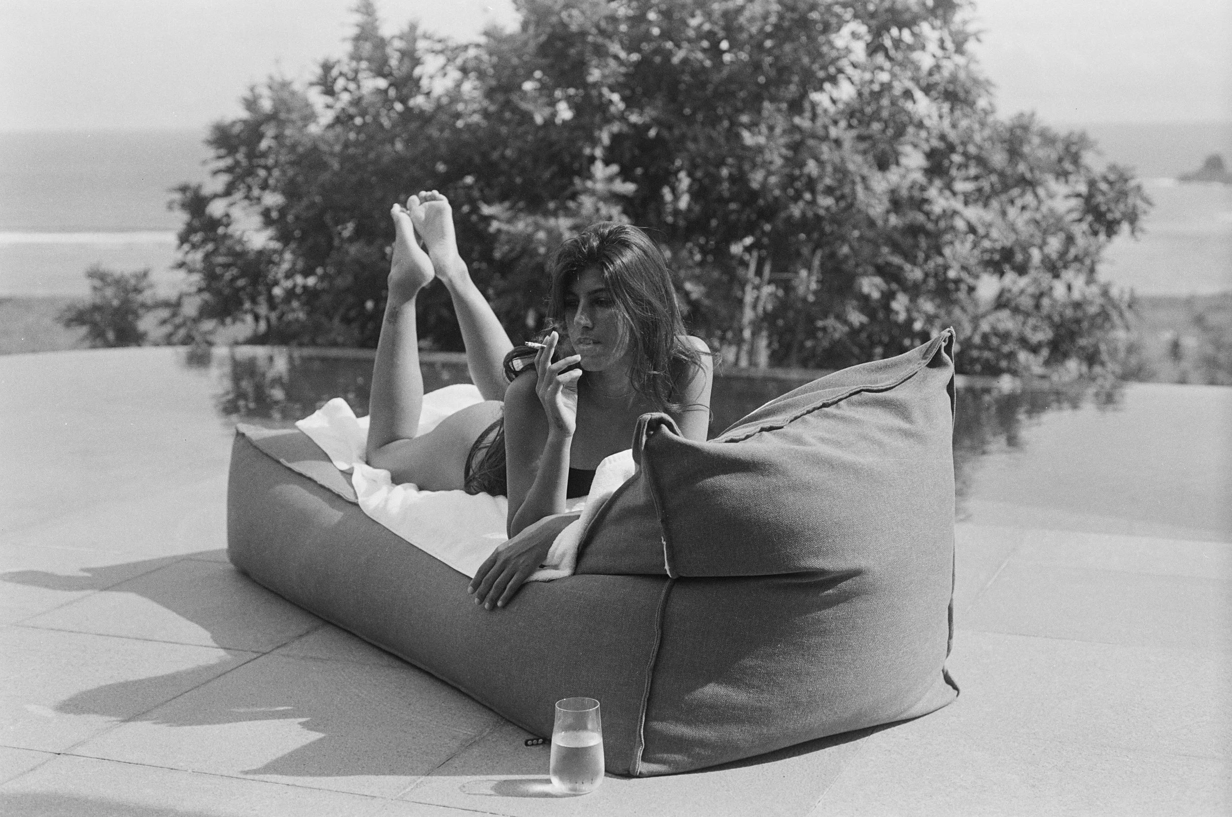 A woman lying on a large outdoor cushion near a pool, holding a cigarette, with a glass of water on the ground in front of her, and a tree in the background.
