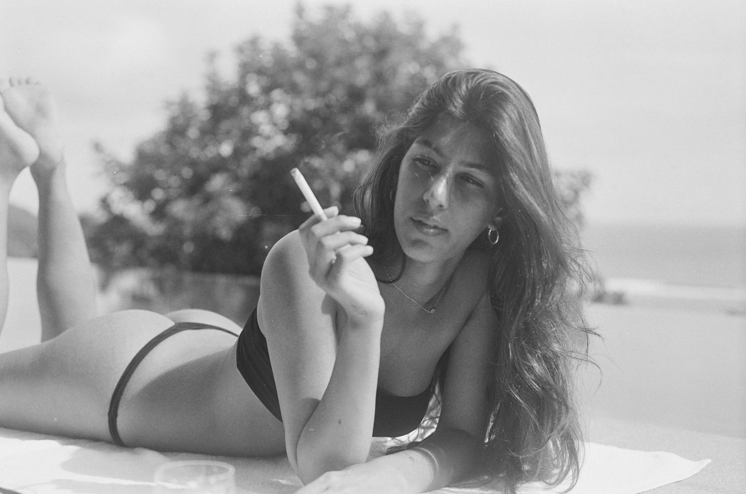 A woman lying on her stomach outdoors, smoking a cigarette, with a tree and ocean in the background.