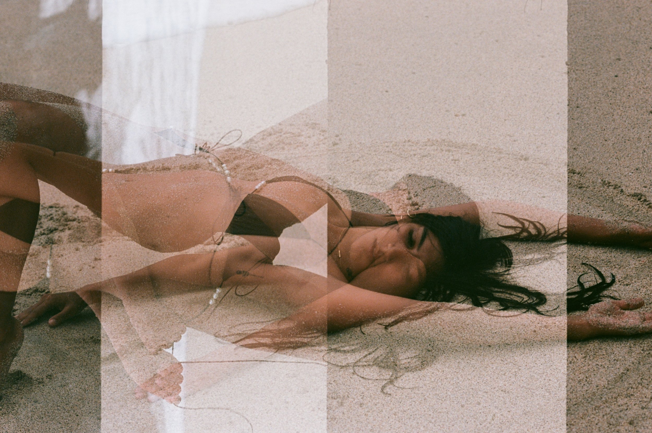 A woman lying on her side on the beach sand, wearing a bikini and jewelry, with her eyes closed.