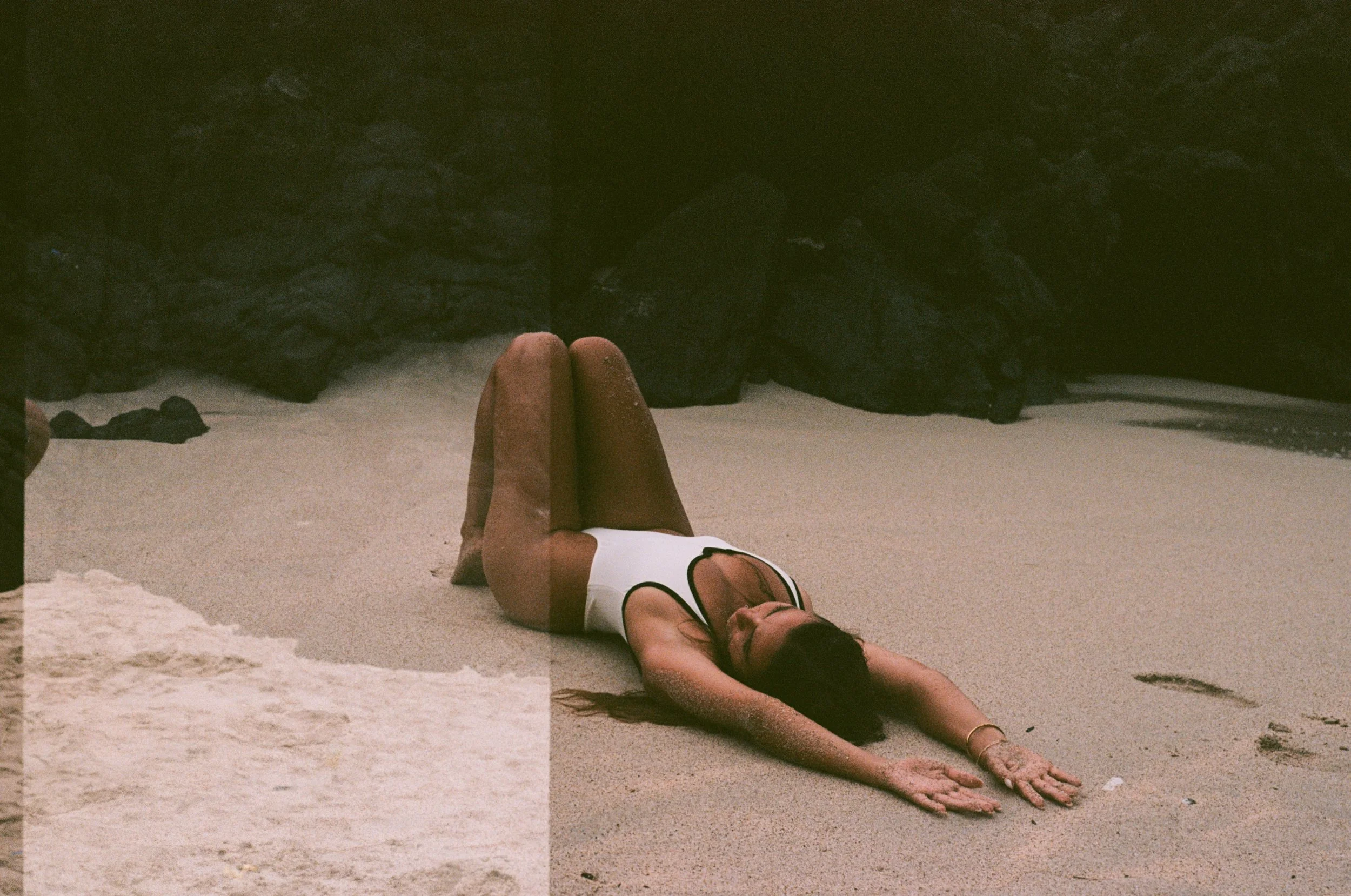 A woman is lying on her back on the sand at the beach, with her arms extended forward and her knees bent. She is wearing a white swimsuit with black straps and has long dark hair.