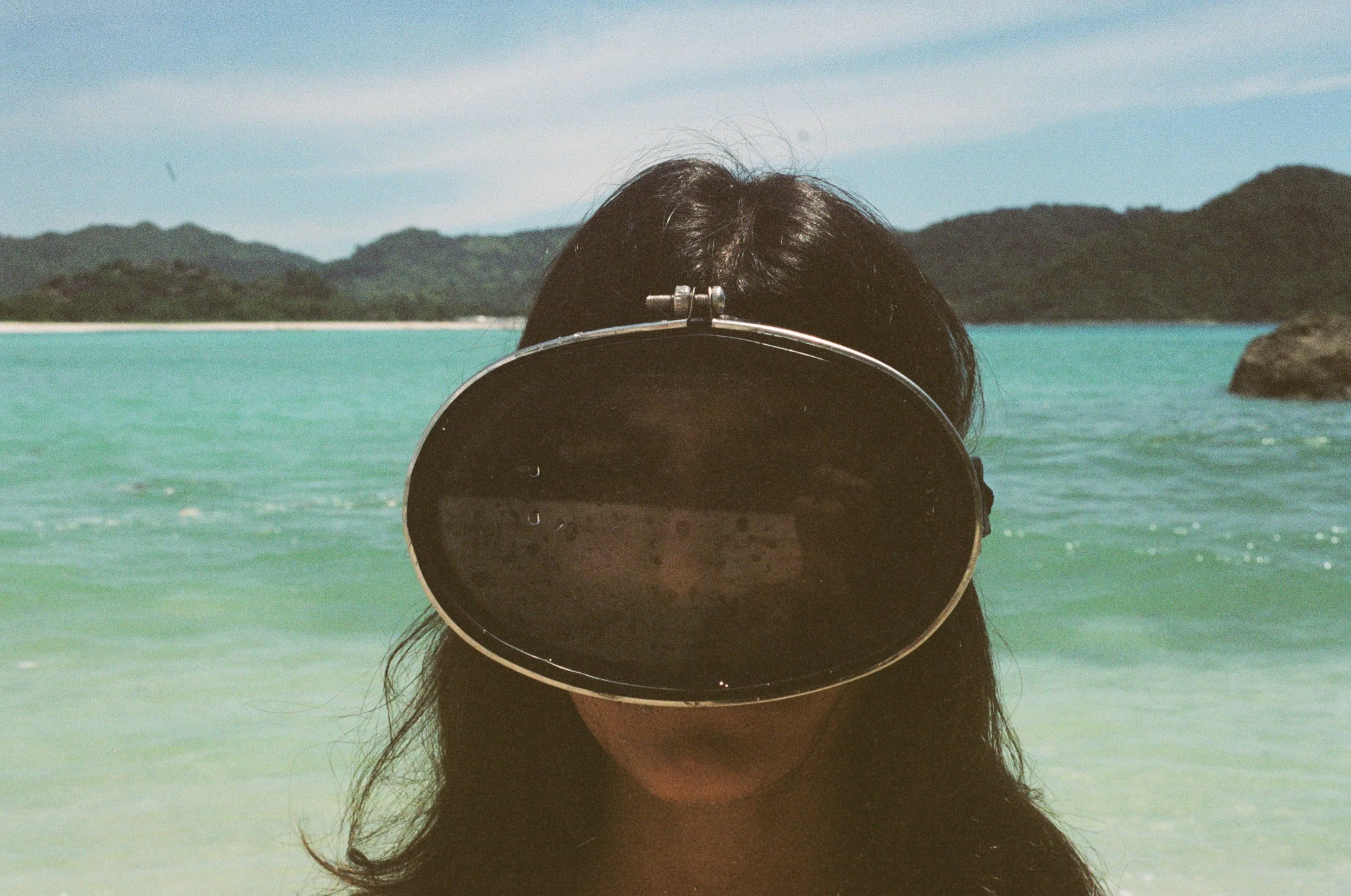 A person standing on a beach with an ocean and mountains in the background, wearing large black sunglasses that cover half of their face.
