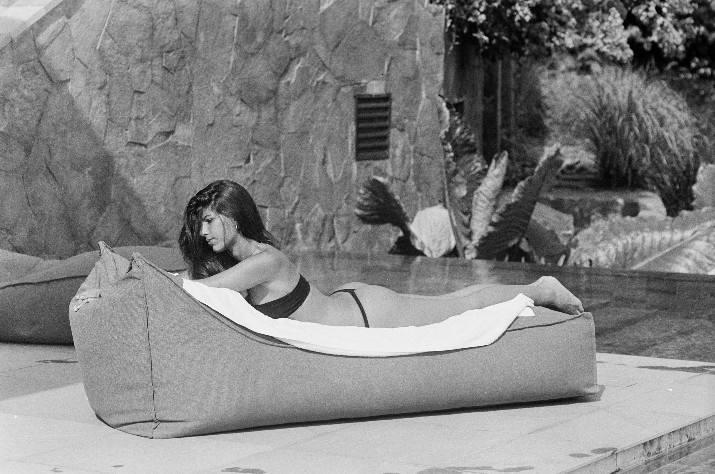 A woman with long dark hair lying on her stomach on a lounge chair by a pool, wearing a bikini top and bottom, with plants and stone wall in the background.