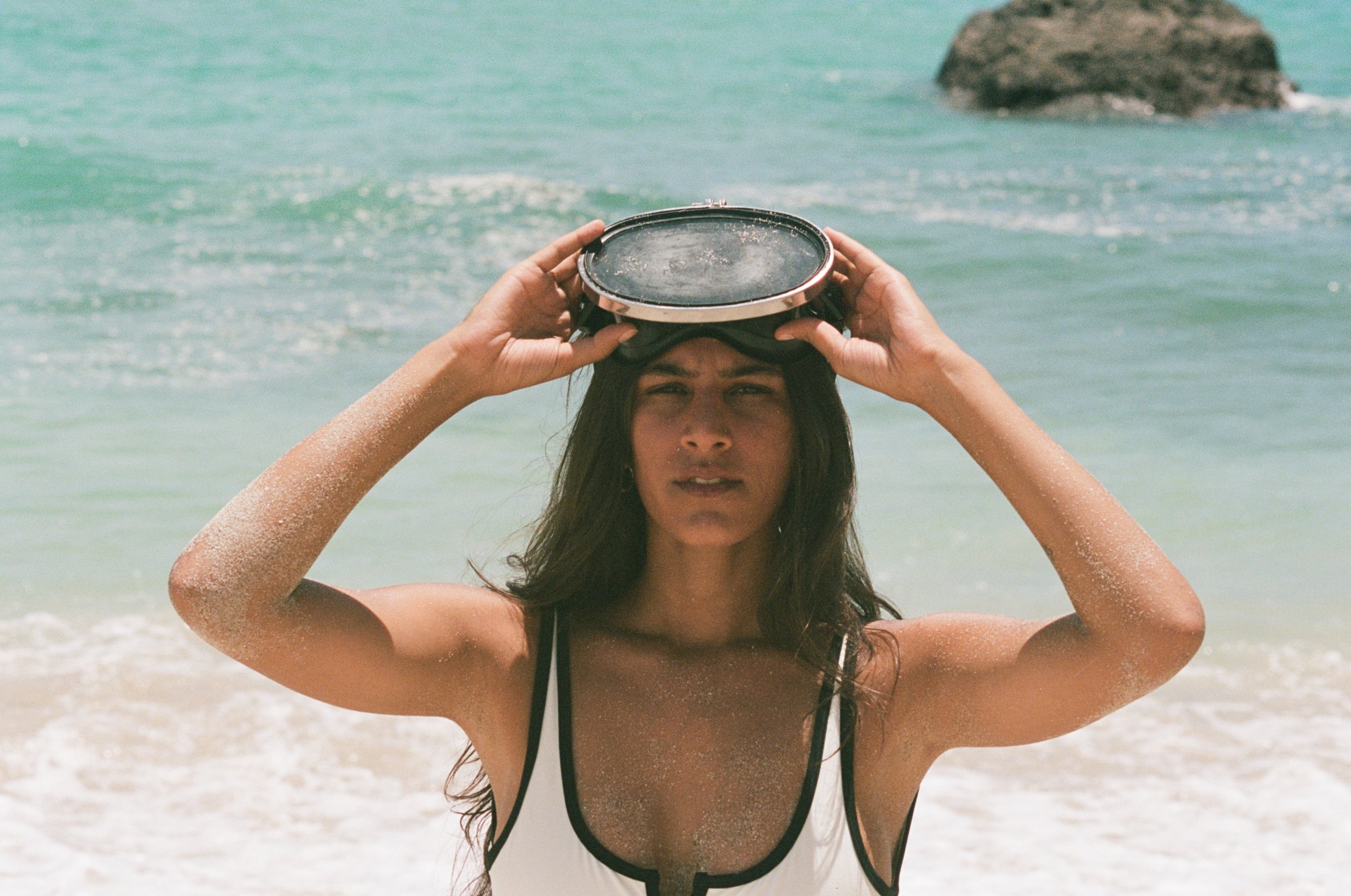 A woman at the beach holding goggles on her head, with sand on her arms and a large rock in the background