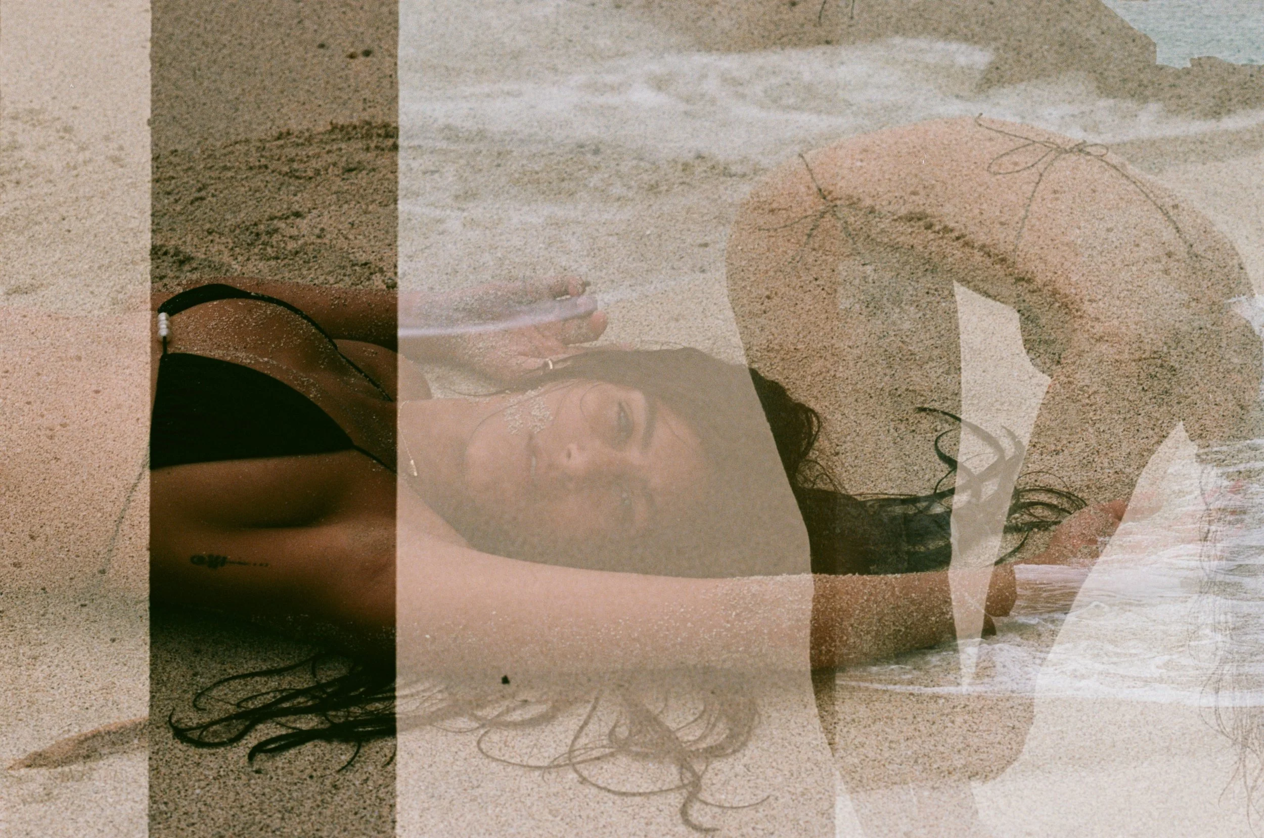 A woman lying on the sandy beach, covered with a translucent overlay that shows her wearing a black bikini, with her hair spread out on the sand and her arm bent over her head.