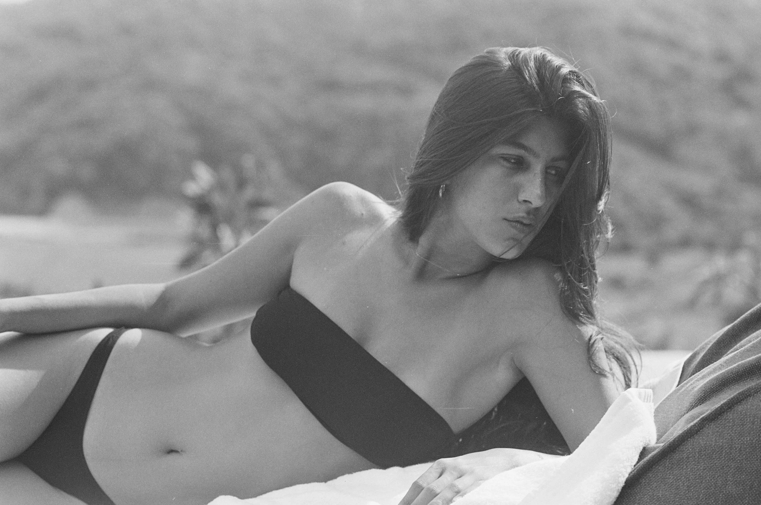 Black and white photograph of a young woman with shoulder-length hair, lying on her side on a bed outdoors, wearing a strapless black top. She has a pensive expression and is looking away from the camera, with a blurred landscape in the background.