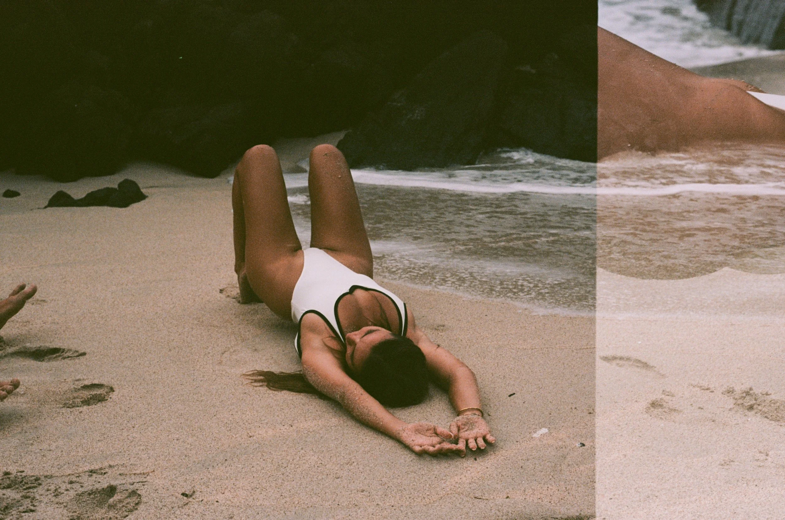 A woman in a black and white swimsuit lying on her stomach on the sandy beach with her head turned sideways and arms stretched out in front of her.