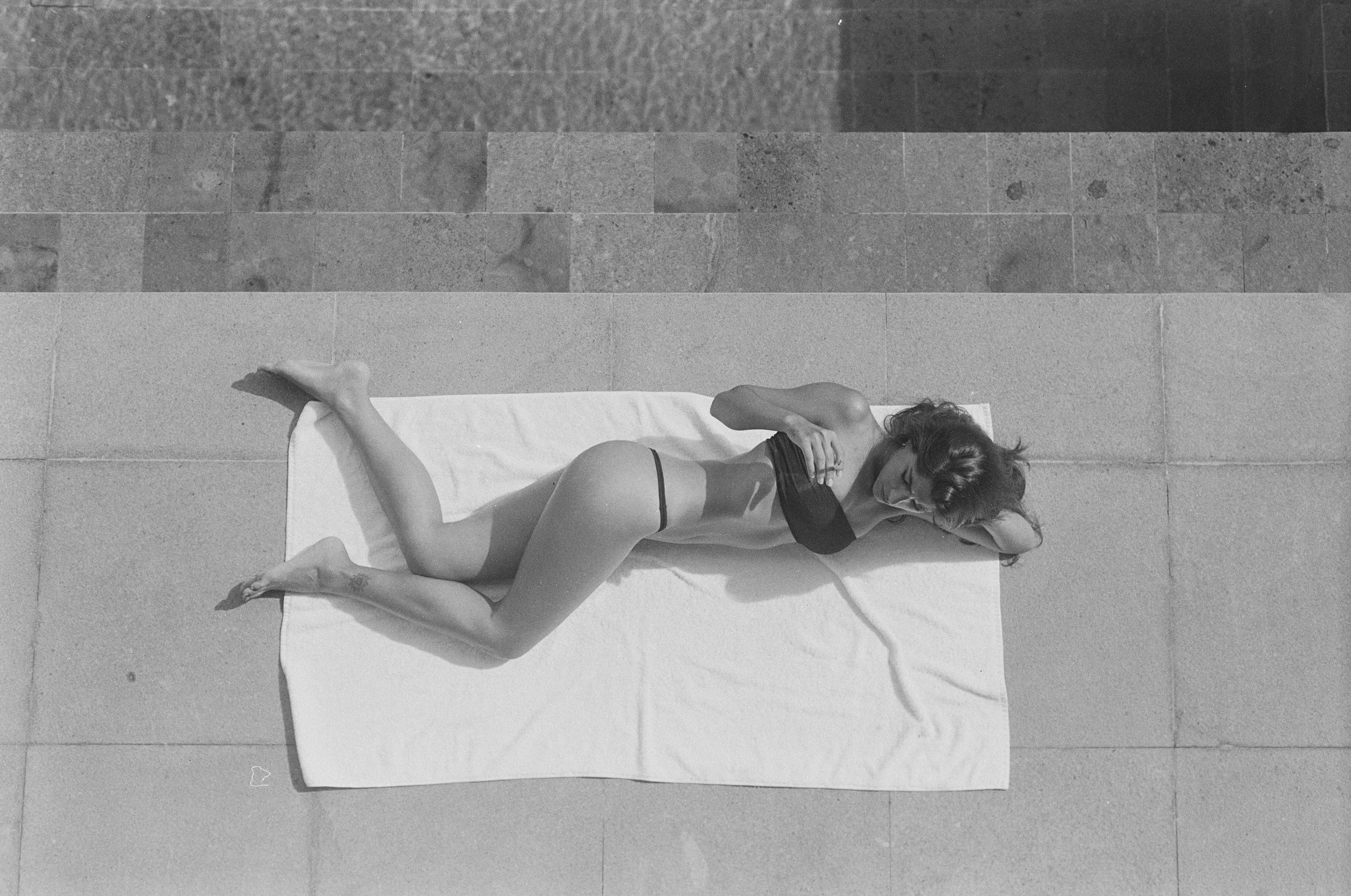 Black and white aerial photo of a woman in black lingerie lying on her side on a white towel on a tiled outdoor surface, with her head resting on her arm and her looking away.