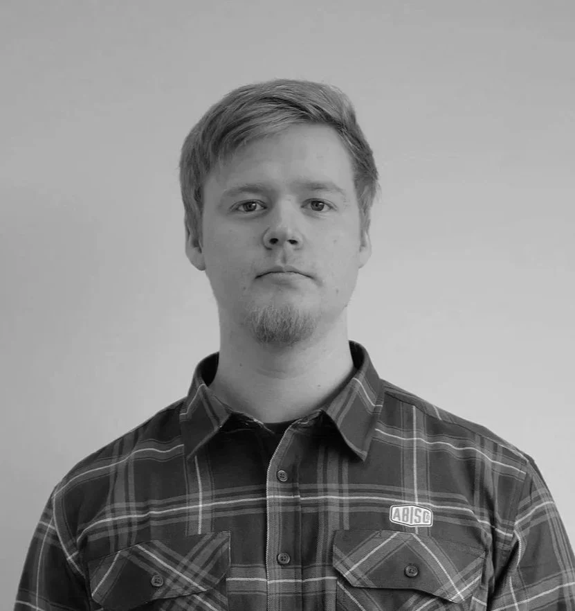 Black and white portrait of a young man with short, light-colored hair and a goatee, wearing a plaid button-up shirt with a small logo on the chest, standing against a plain background.